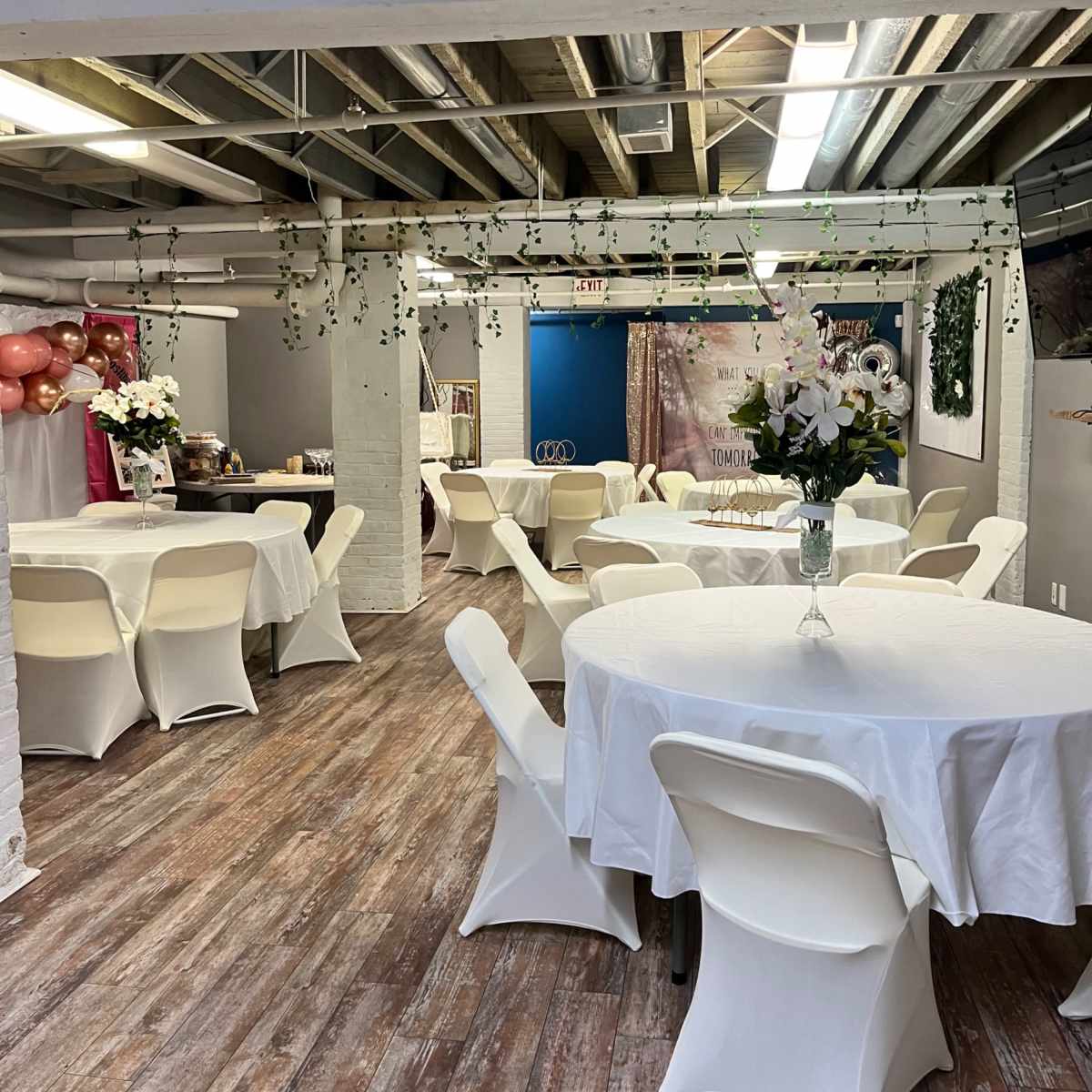 A spacious event room with several round tables covered in white tablecloths, decorated with vases of flowers, and walls adorned with balloons and streamers.