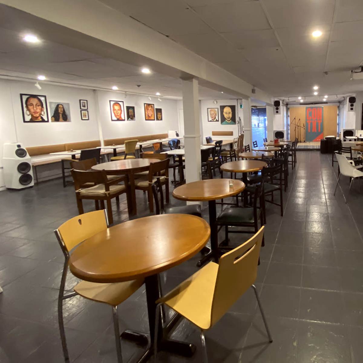 A spacious indoor café or event space with tables and chairs arranged around the room, featuring artwork on the walls.