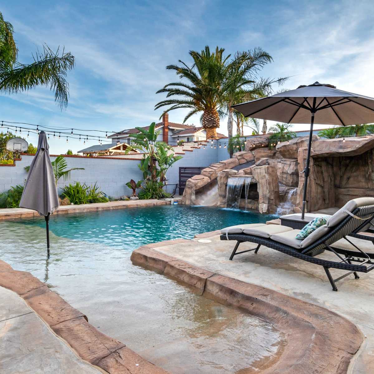 A backyard pool with a rocky waterfall, surrounded by palm trees and lounge chairs under an umbrella.