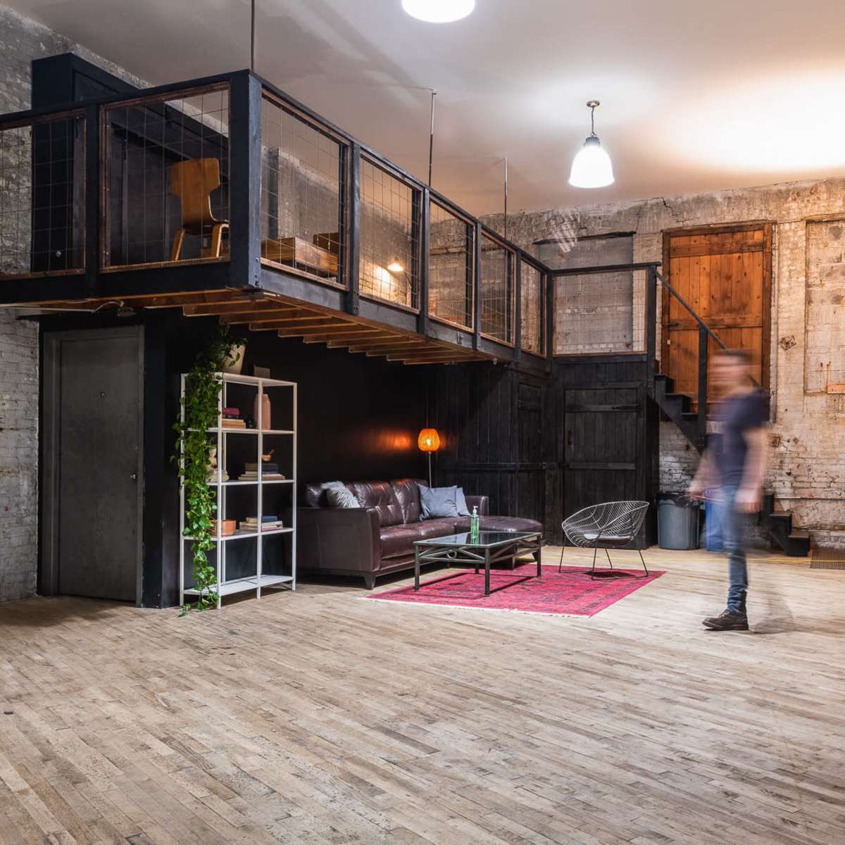 An industrial-style loft space with a wooden floor, a raised loft area, a modern seating area, and a mannequin near a bookshelf.