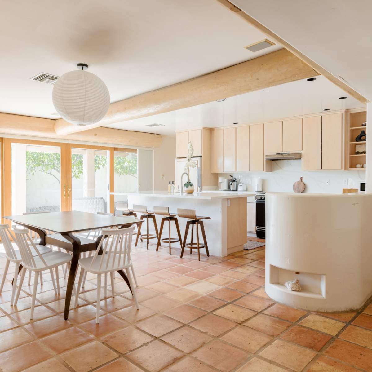 A spacious kitchen and dining area features a table with white chairs, light wood cabinets, and large sliding glass doors leading to a garden.