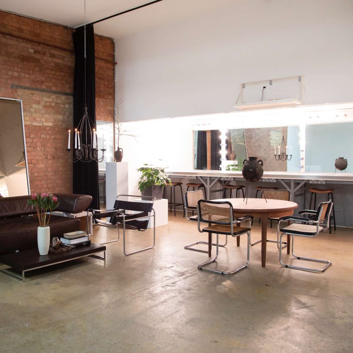 The scene features a spacious room with exposed brick walls, a brown leather sofa, a round wooden table surrounded by modern chairs, and a large mirror reflecting the minimalist decor.