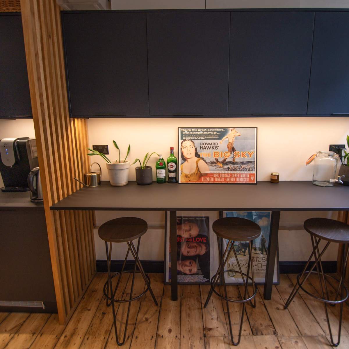 A modern kitchen with a long, dark countertop, three bar stools, and a coffee machine on the side, featuring a framed movie poster on the wall.
