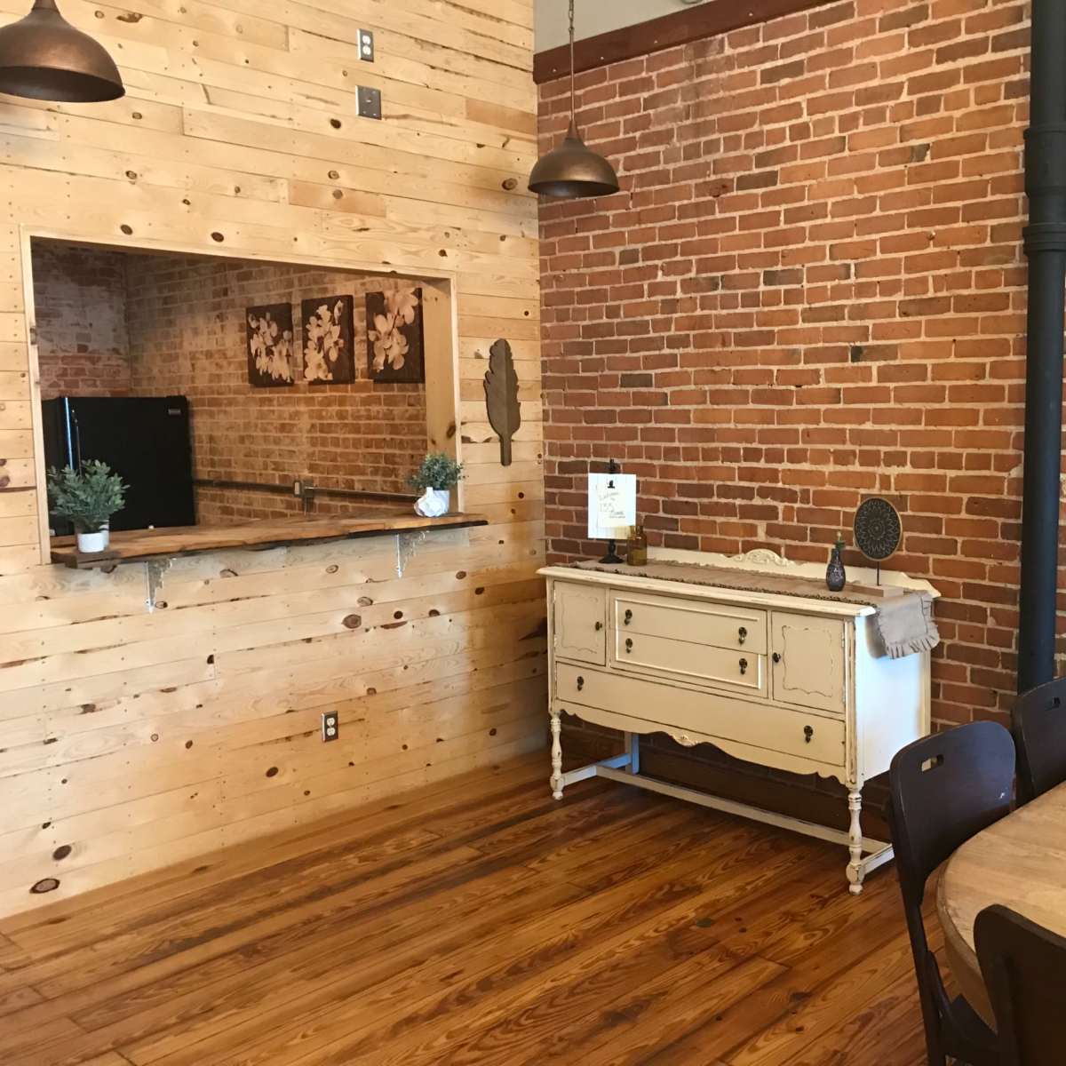 A dining area with a wooden table and chairs, adjacent to a kitchen area featuring a brick wall and a light-colored sideboard.
