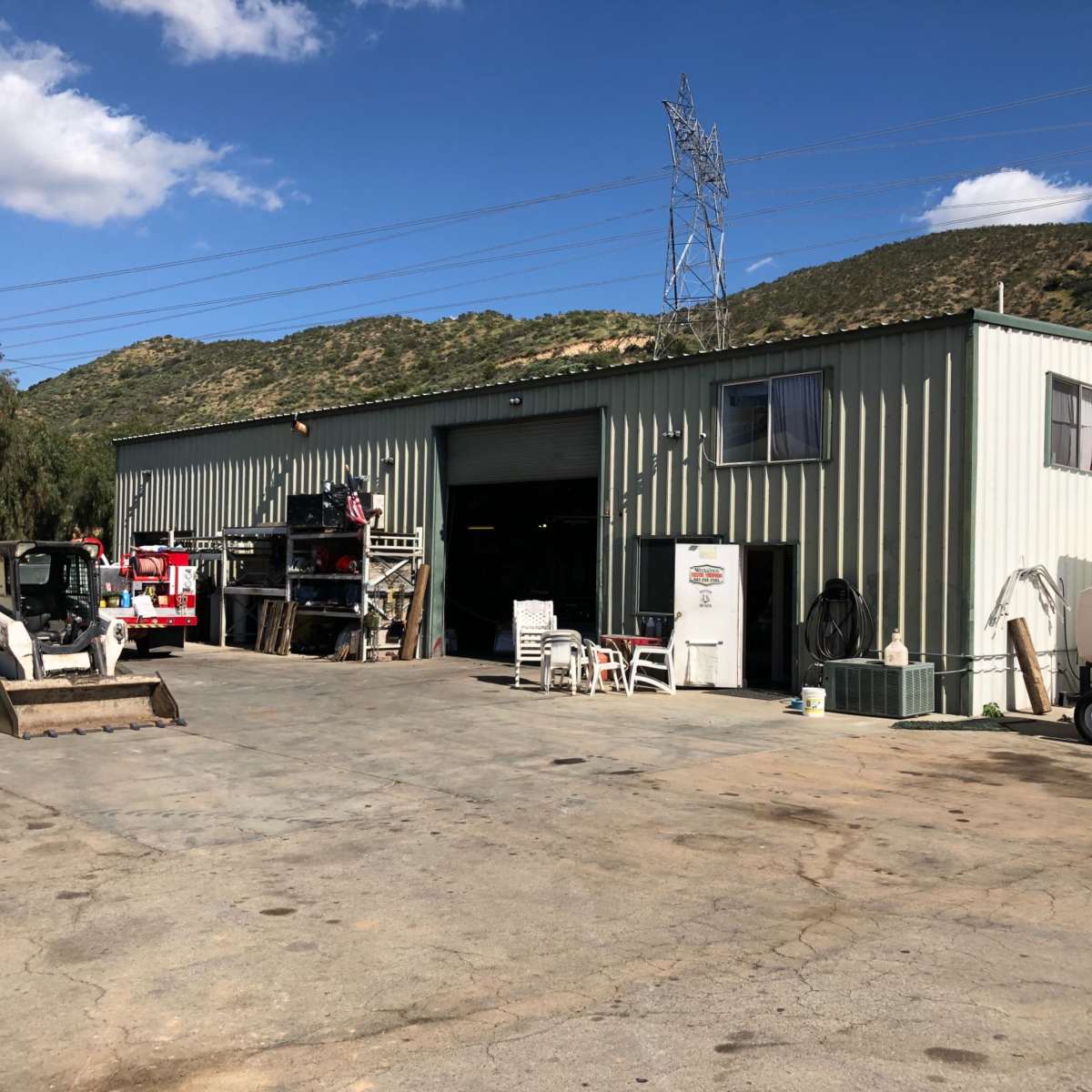 A metal building with an open garage door stands in an outdoor area surrounded by machinery and equipment.