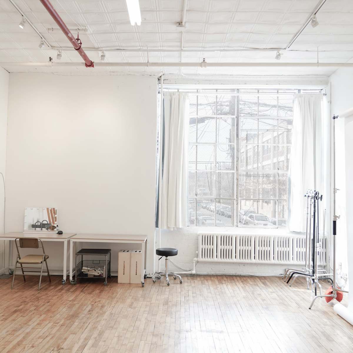 A spacious, well-lit studio with large windows, a wooden floor, an easel, a desk with chairs, and a backdrop setup for photography.