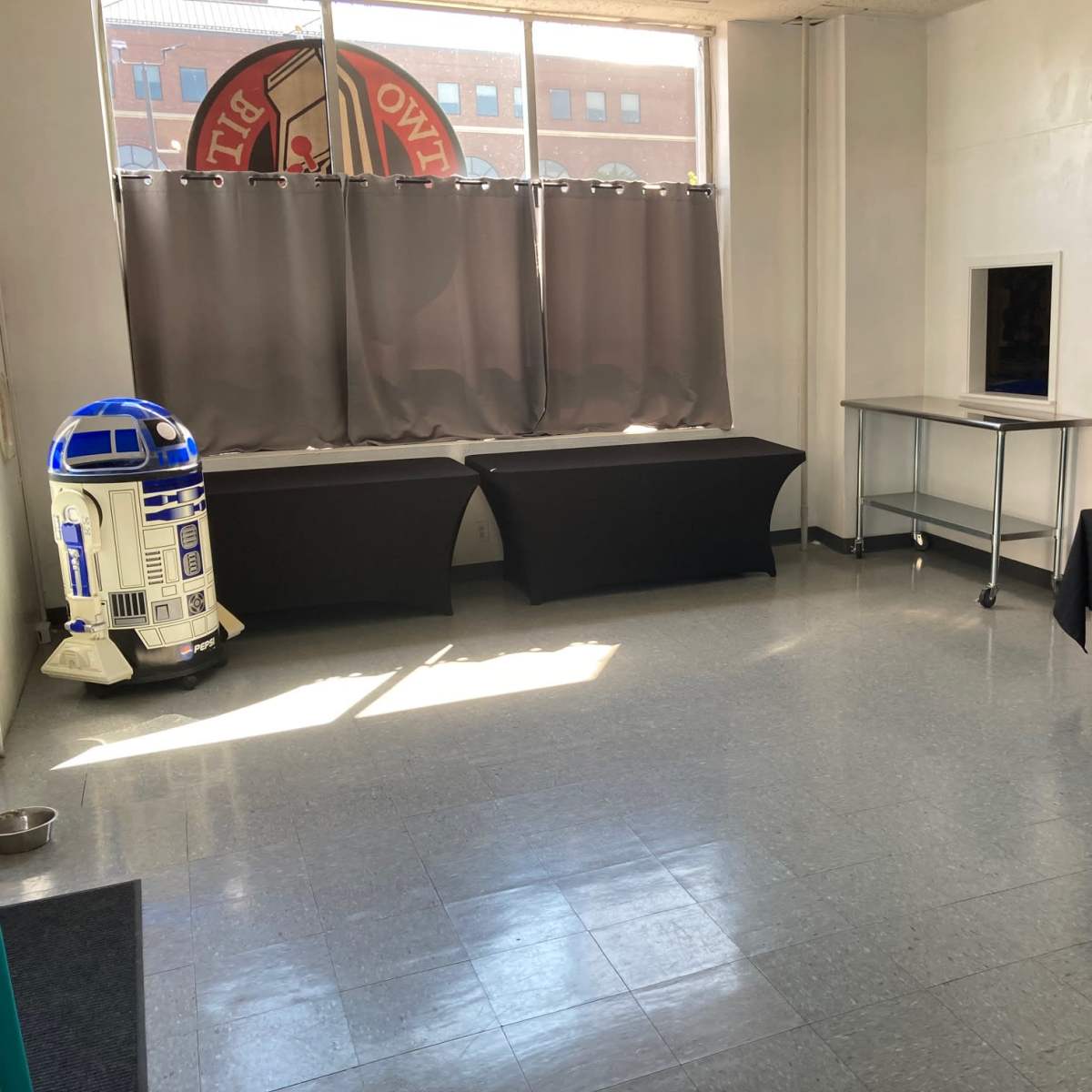 An empty room with gray tile flooring, two tablecloth-covered tables, a metal table, and a decorative R2-D2 figure in the corner.