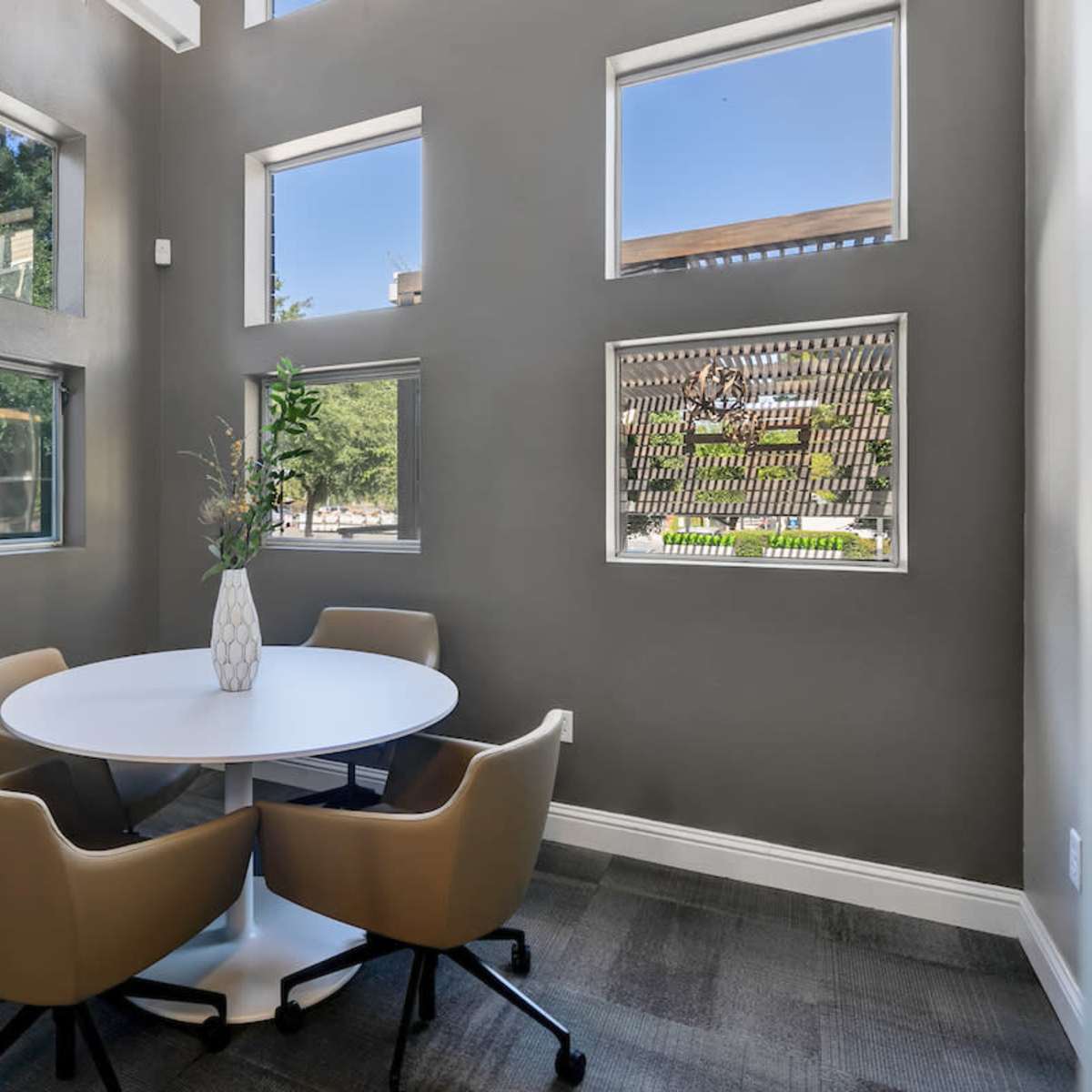 A small meeting room features a round table with four beige chairs, surrounded by large windows letting in natural light.
