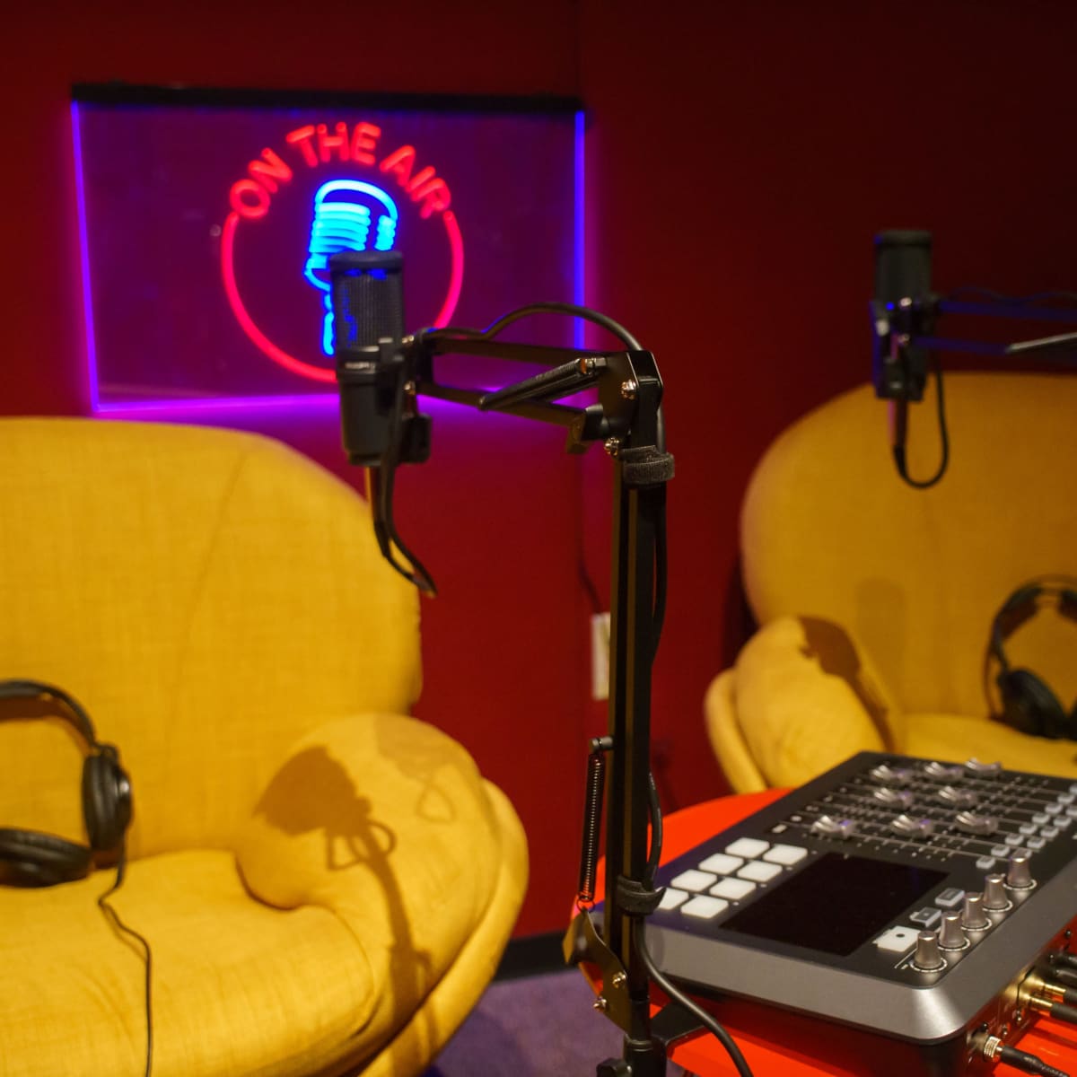 A radio studio setup with two yellow armchairs, a sound mixer on a red table, and a neon sign that reads "ON THE AIR."