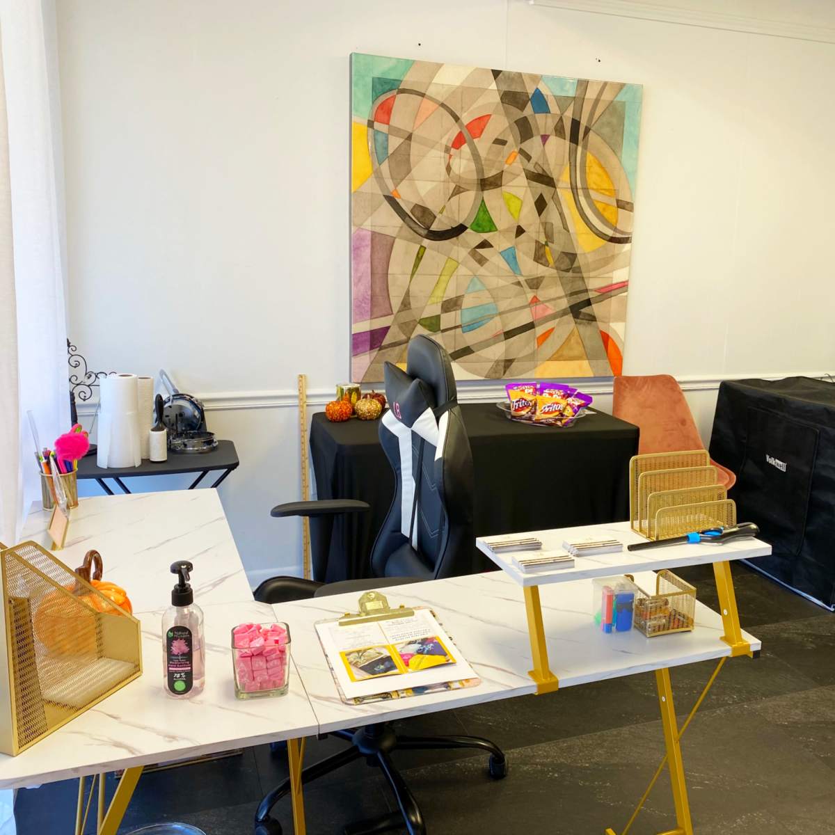 A well-organized room featuring a modern desk with a chair, a large abstract painting on the wall, and various items displayed on a black table and a grid panel.