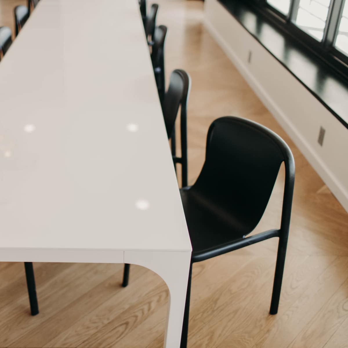 A long white table with black chairs surrounding it is placed on a wooden floor in a well-lit room.