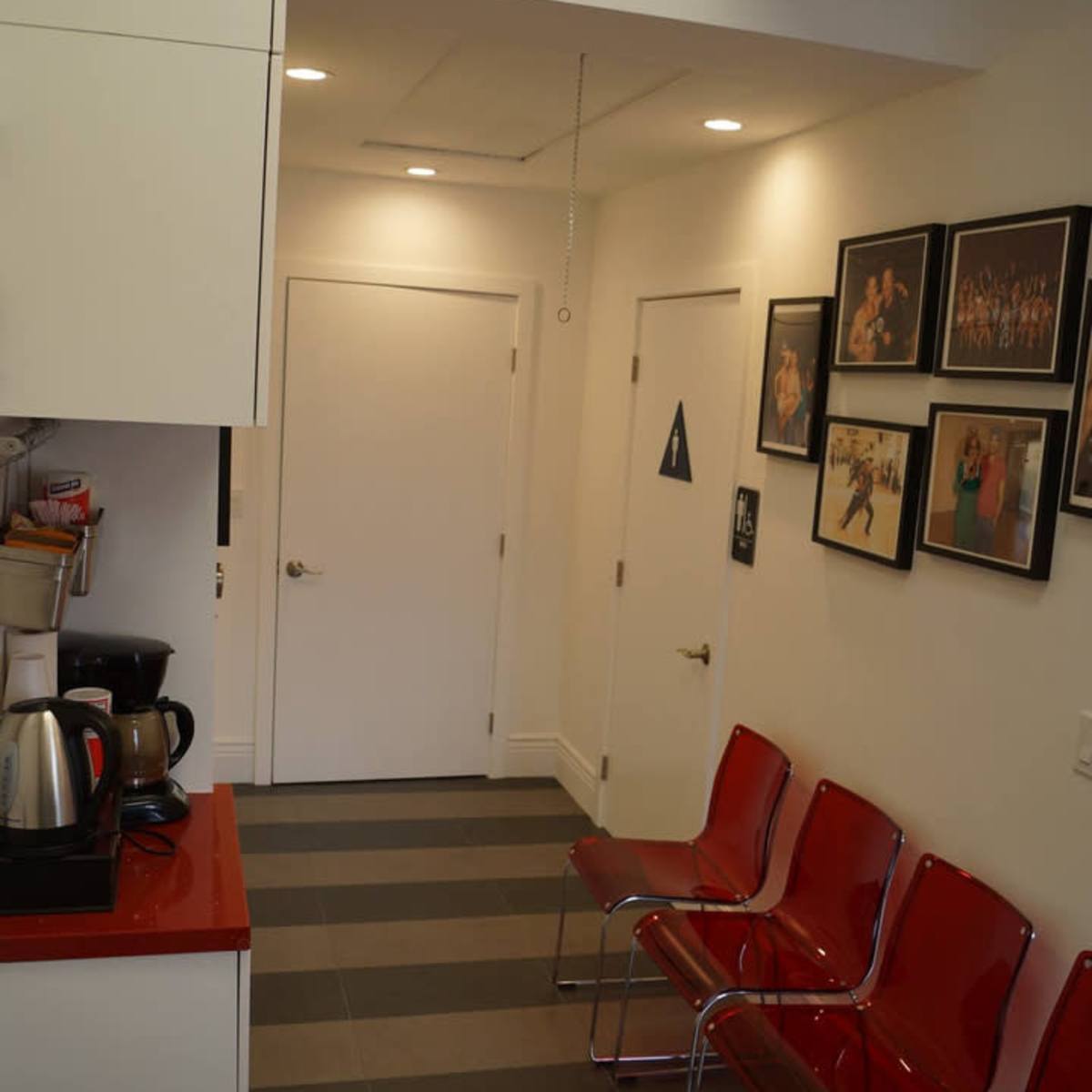 A narrow hallway with a kitchenette on the left, a door at the end, and several red chairs along the right wall, which is decorated with framed photos.