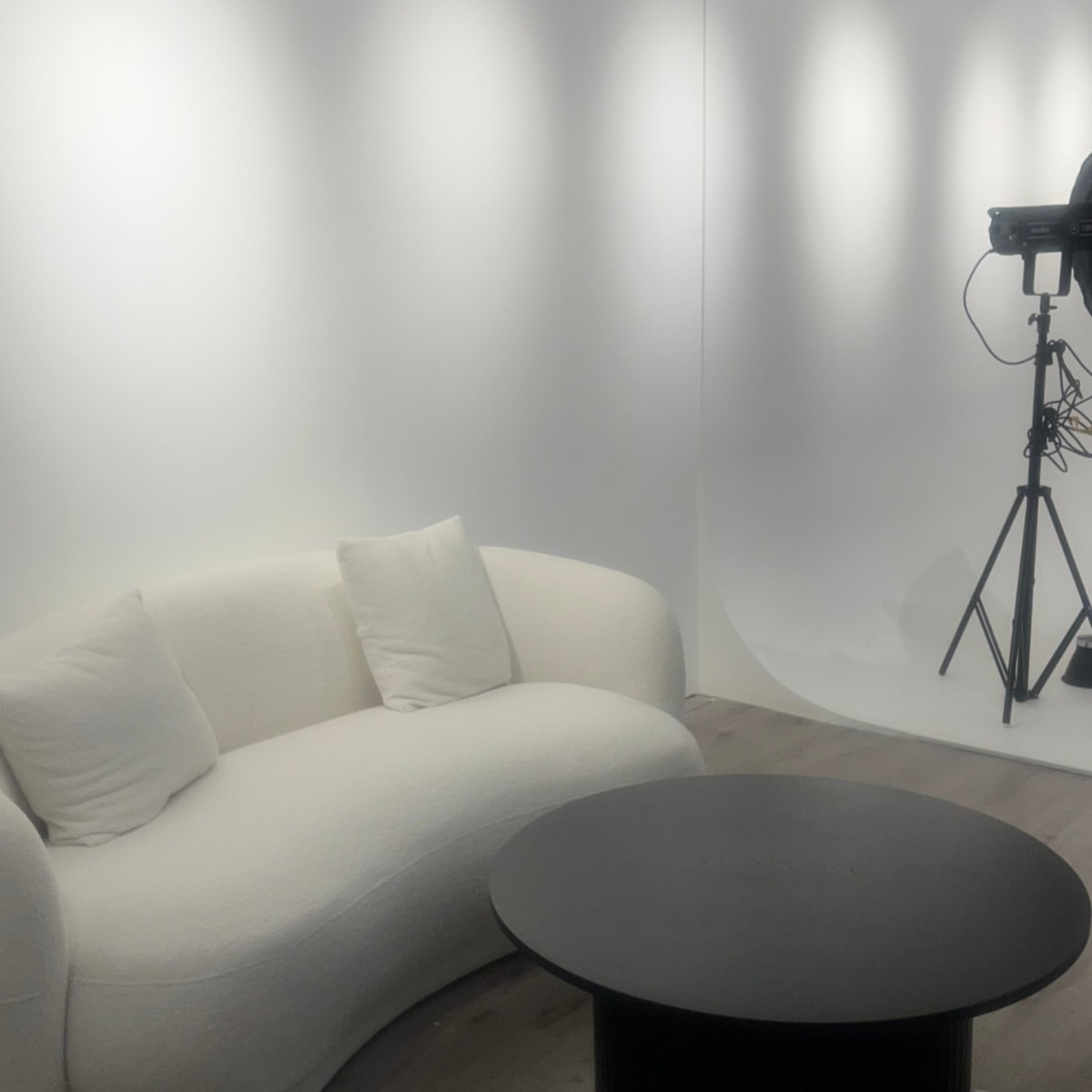 A minimalist studio space featuring a curved white sofa and a round black table, with studio lighting equipment in the background.