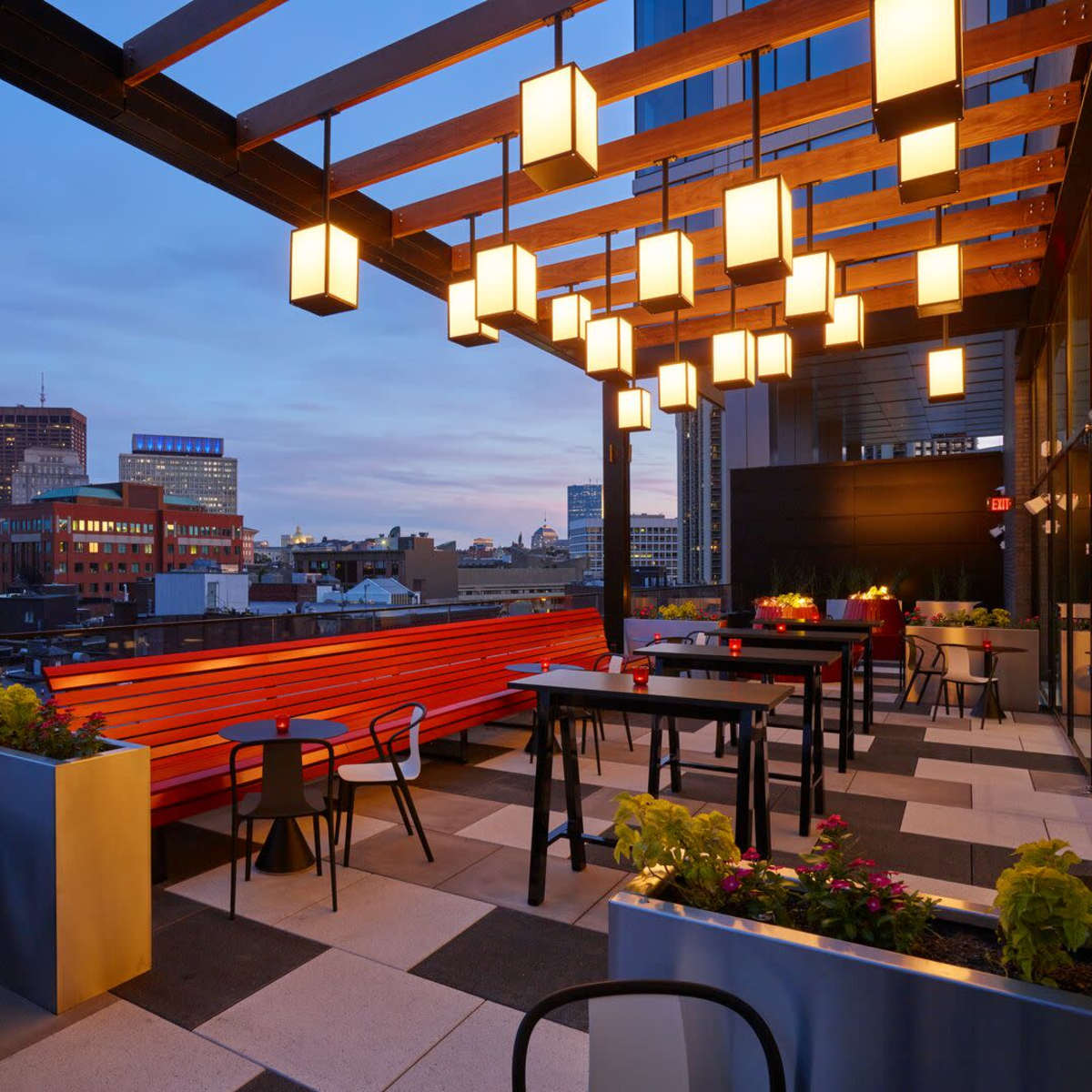 A modern rooftop dining area features wooden beams with hanging lanterns, surrounded by planters and a city skyline in the background during twilight.