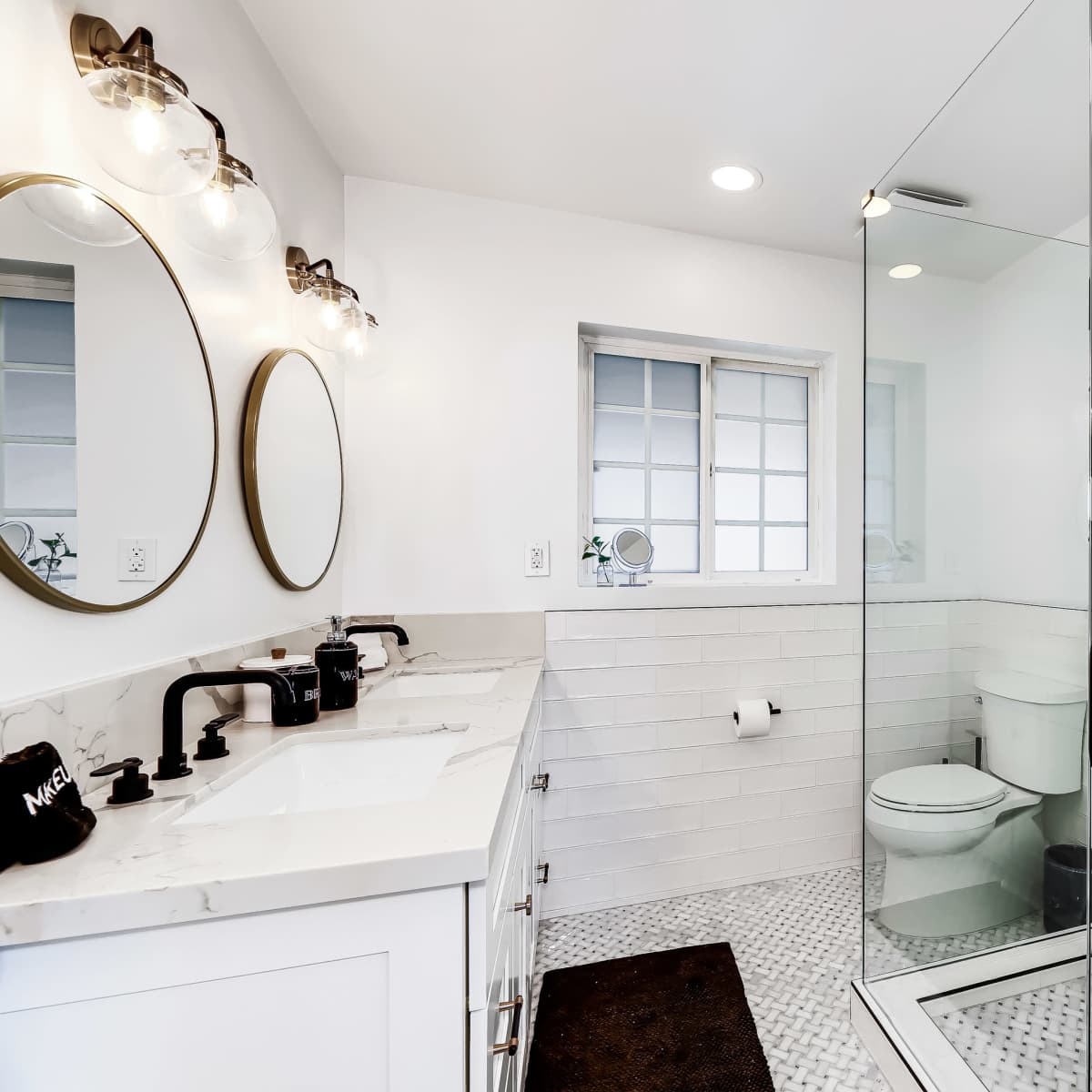 A modern bathroom features a walk-in shower, dual sinks with round mirrors, and white tiled walls.