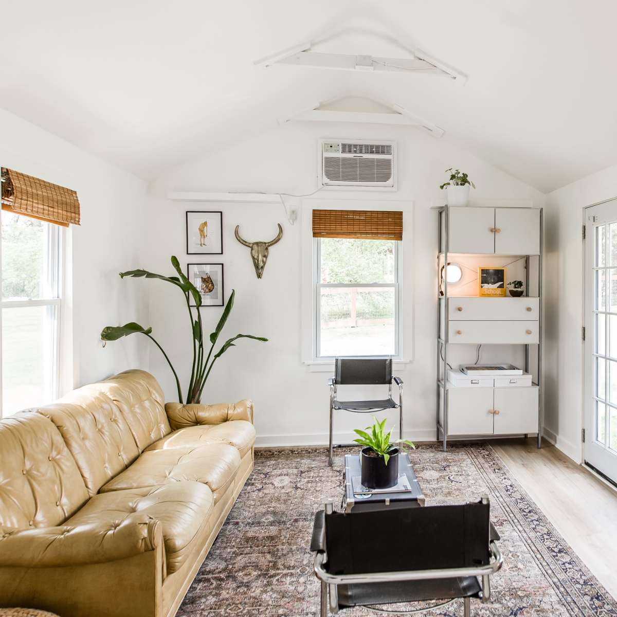 A bright, small living room featuring a tan leather sofa, a decorative rug, a wooden cabinet, and large windows with light filtering through.