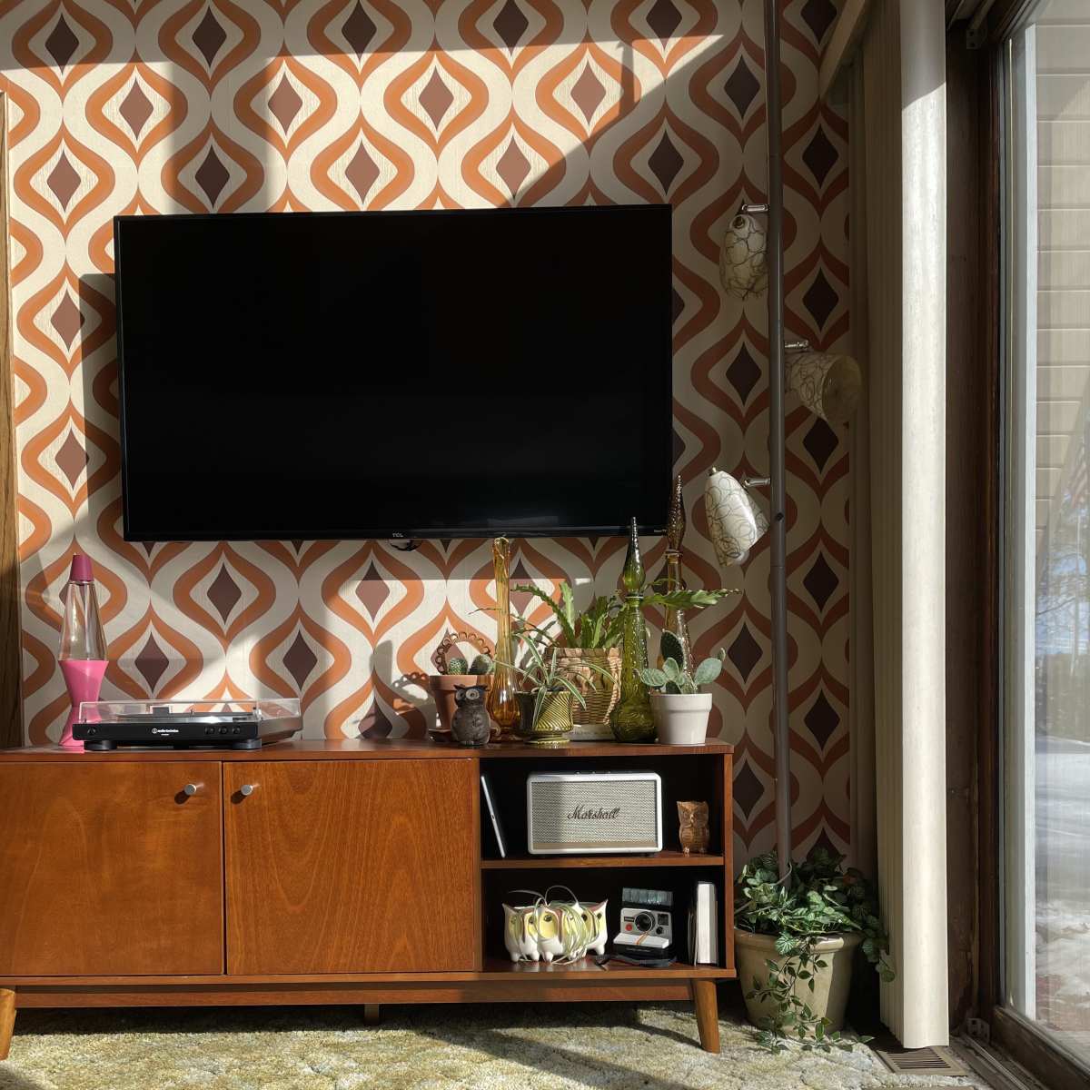 A wooden media console with a television above it sits against a patterned wall, surrounded by plants and decorative items, on a carpeted floor.