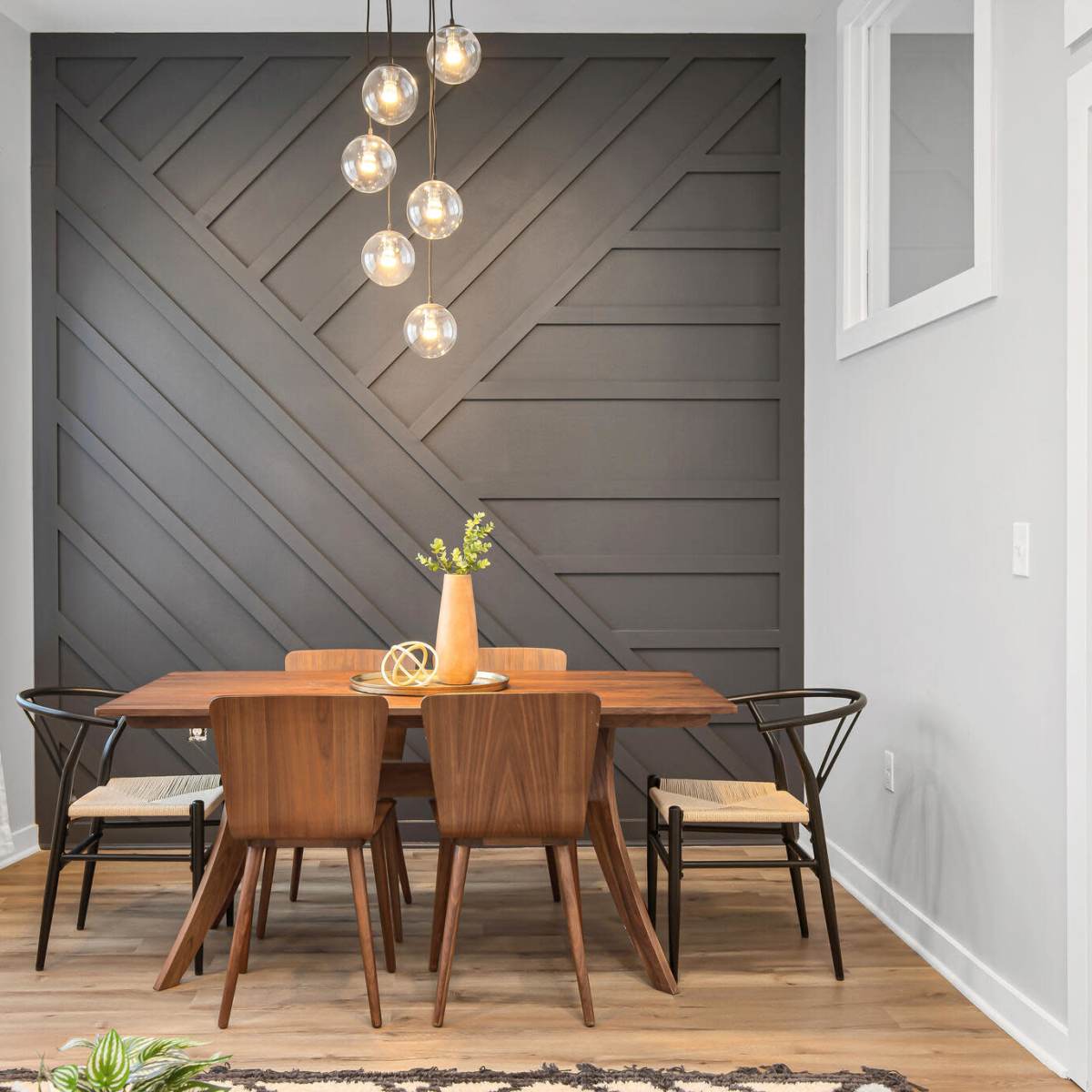 A modern dining area features a wooden table surrounded by chairs, with a accent wall displaying a geometric design and pendant lights above.