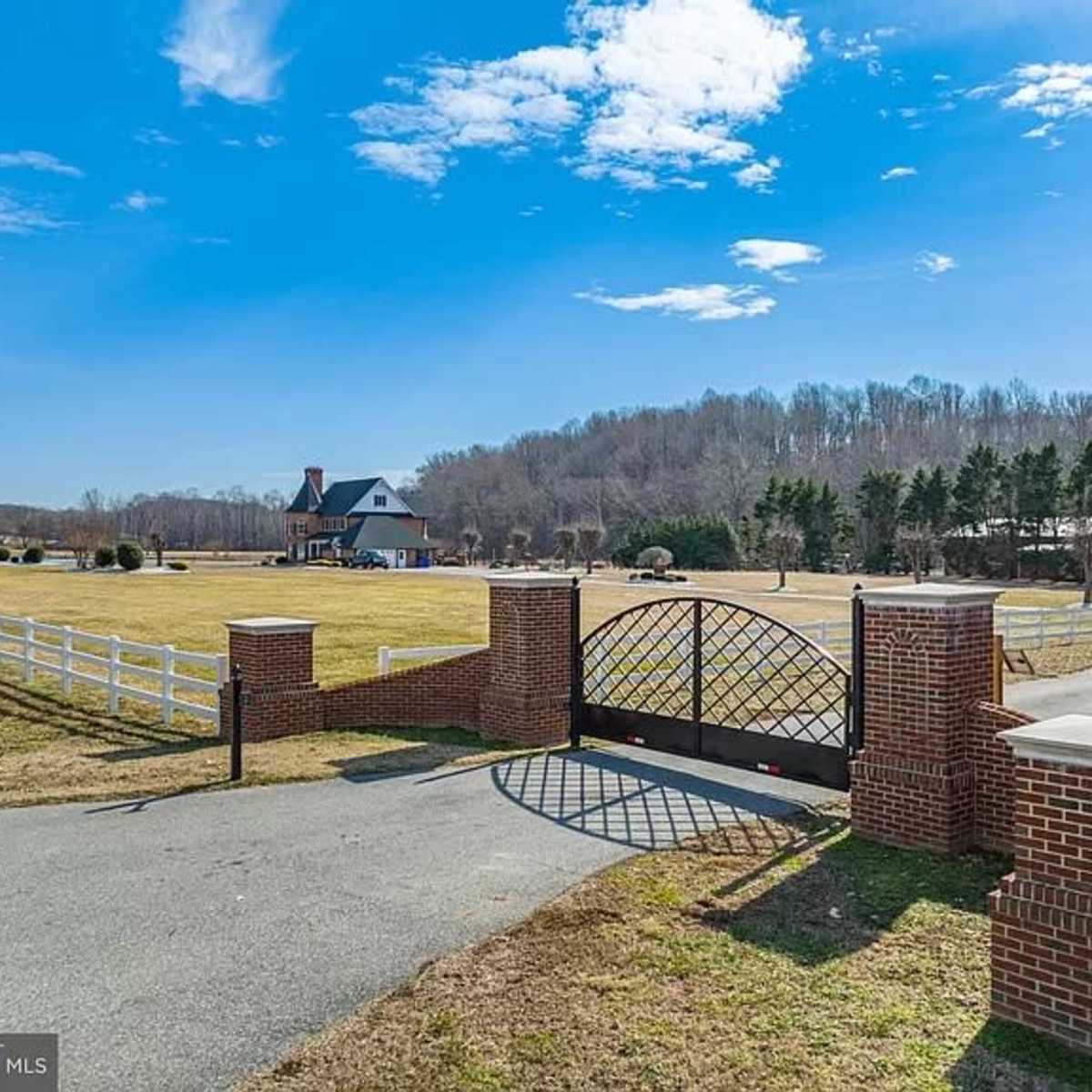 A large brick gate with a wrought iron design opens to a paved driveway leading to a sizable house, surrounded by green fields and trees.