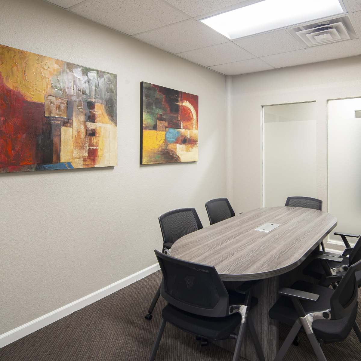A conference room features a large oval table surrounded by black chairs, with abstract paintings on the walls and glass-paneled doors.
