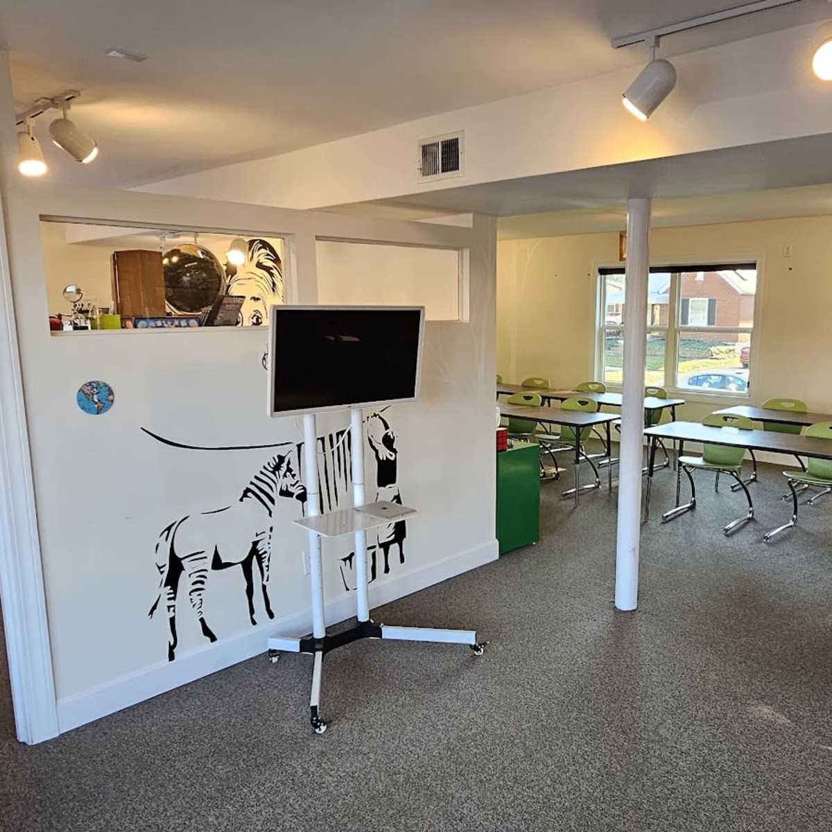 A well-lit classroom features green chairs and tables arranged for learning, with a wall mural of zebras and another room visible in the background.