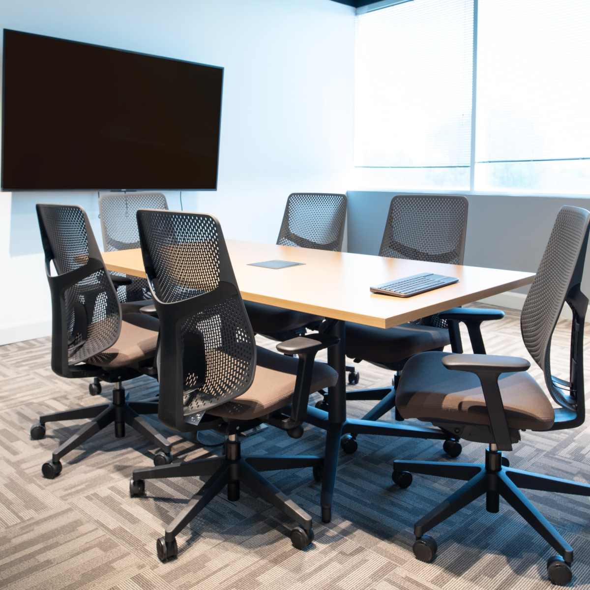 A conference room is arranged with a rectangular table surrounded by six ergonomic chairs, and a large screen mounted on the wall.
