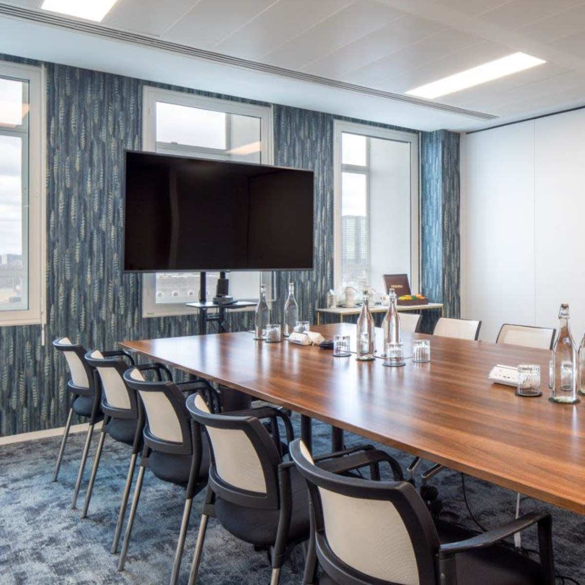 A modern conference room features a large wooden table surrounded by black chairs, equipped with a television for presentations and large windows letting in natural light.