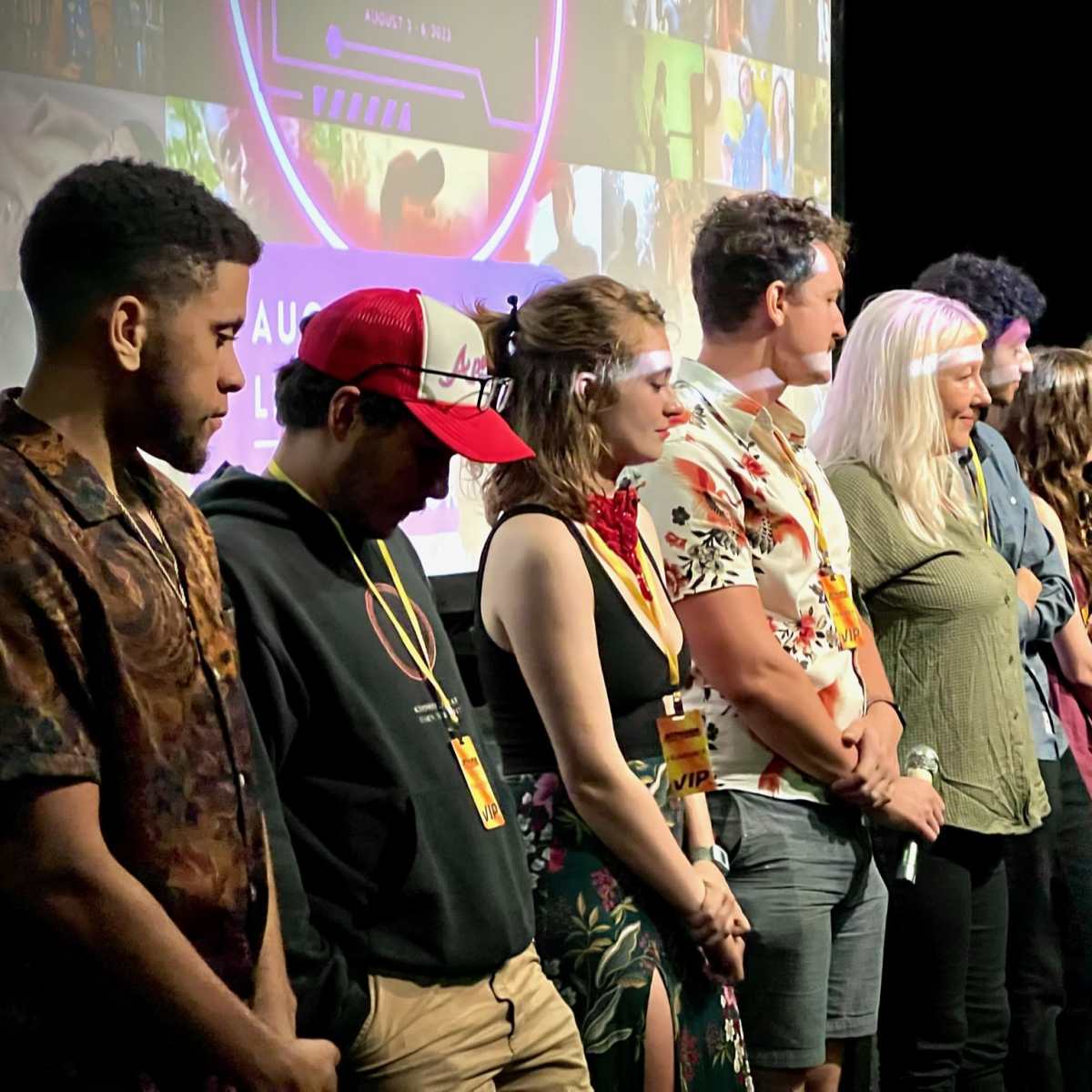 A group of ten people stands in a line on stage, facing an audience, with a colorful backdrop featuring images and text.