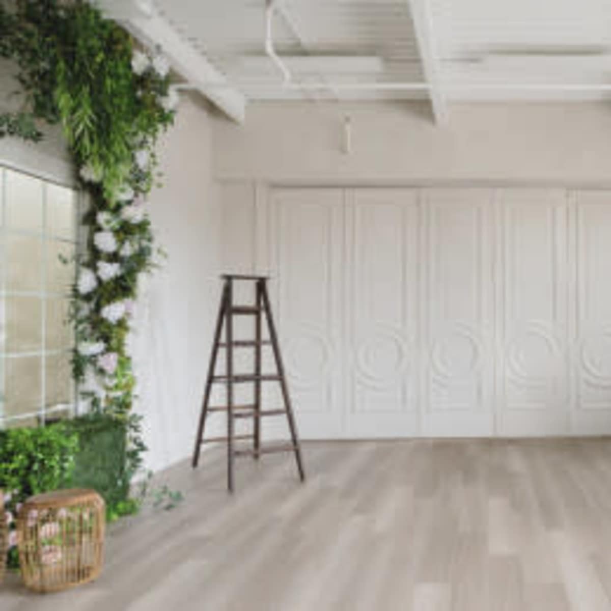 A spacious indoor room features light wood flooring, a white wall with decorative panels, and greenery along the edges, with a wooden ladder and lanterns placed decoratively.