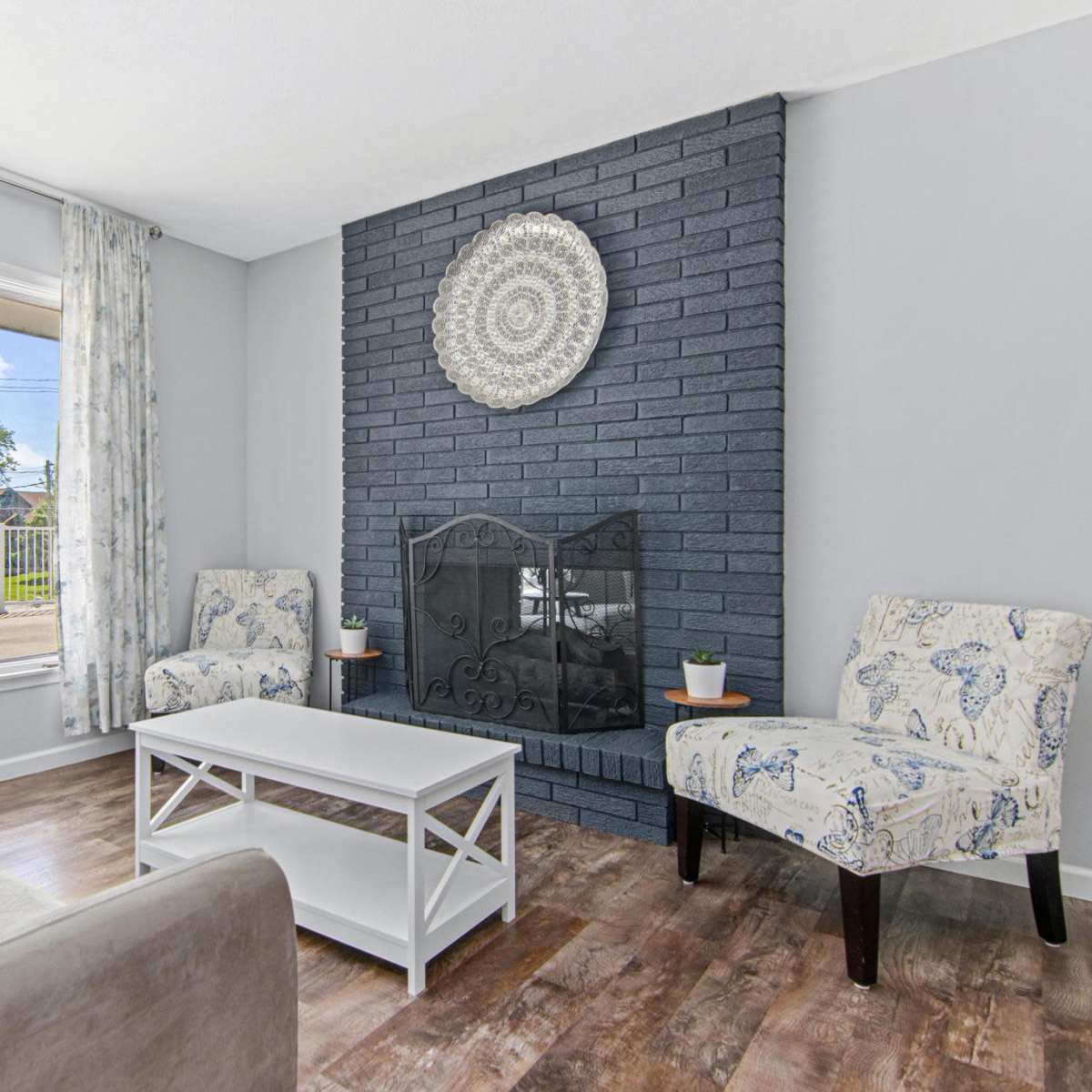 The room features a light-colored sofa and two patterned chairs facing a black-brick fireplace, with a window overlooking an outdoor scene.