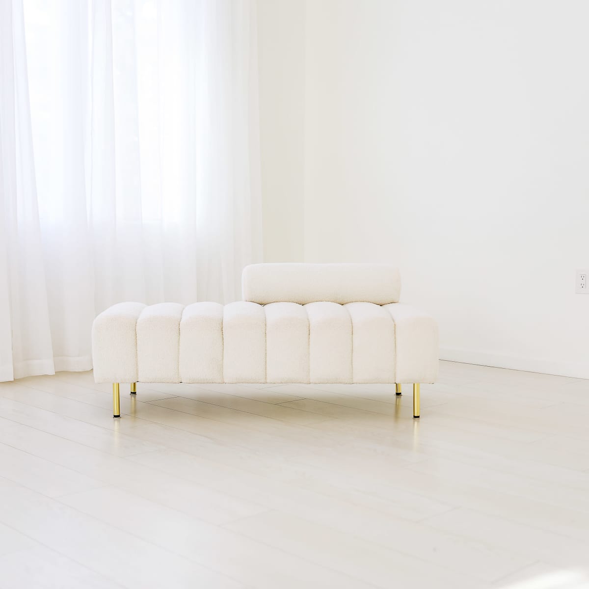 A light-colored, tufted chaise lounge positioned in a bright, minimally decorated room with large windows.