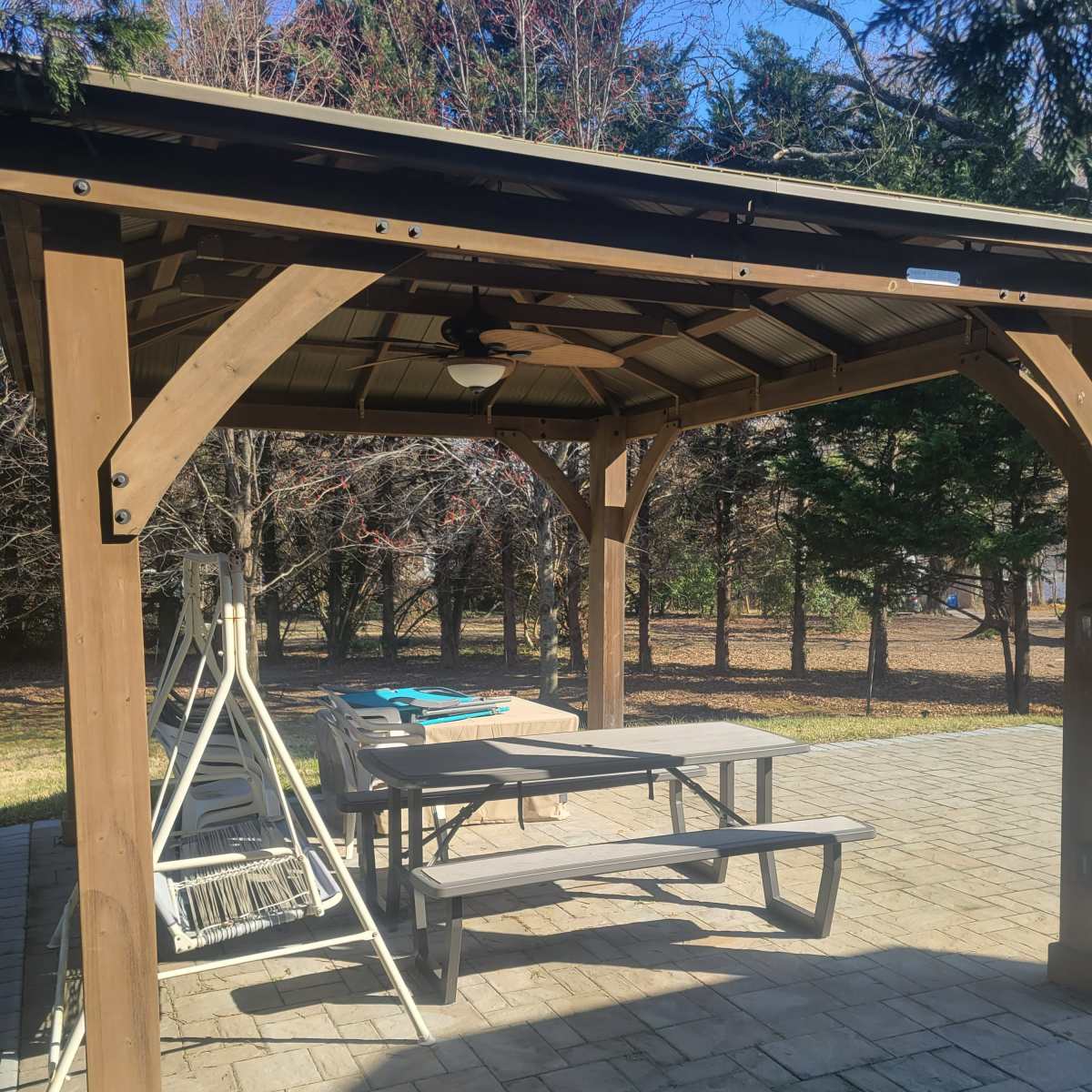 A wooden gazebo with a picnic table is set up on a paved patio overlooking a grassy area and trees in the background.