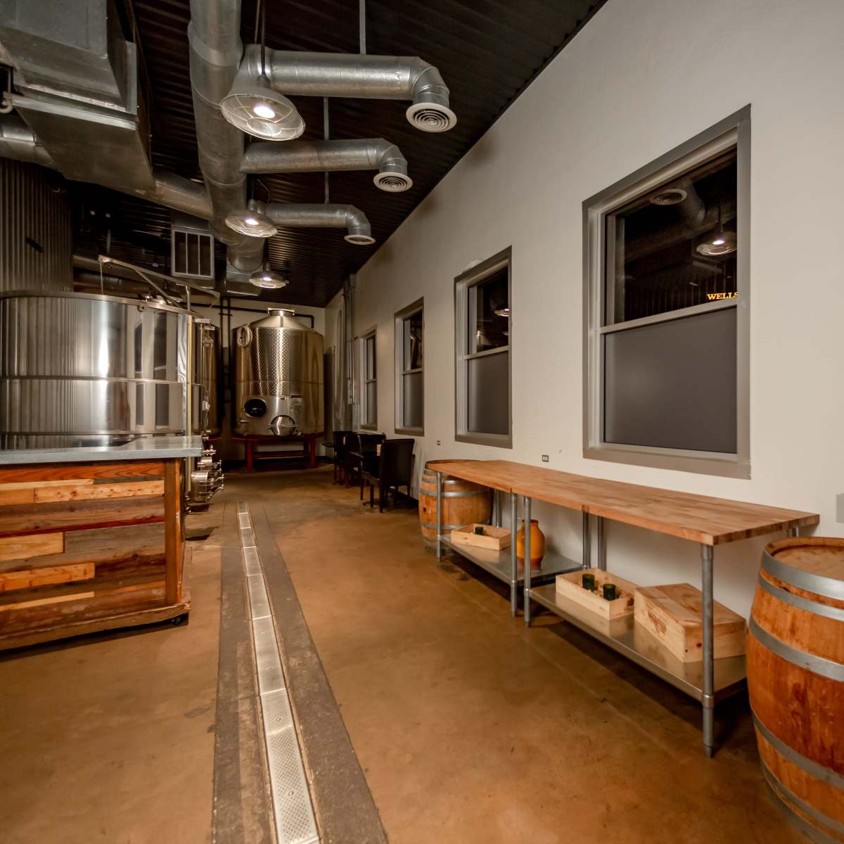 A narrow, industrial-style brewery interior with stainless steel tanks, wooden barrels, and a long wooden table along one wall.