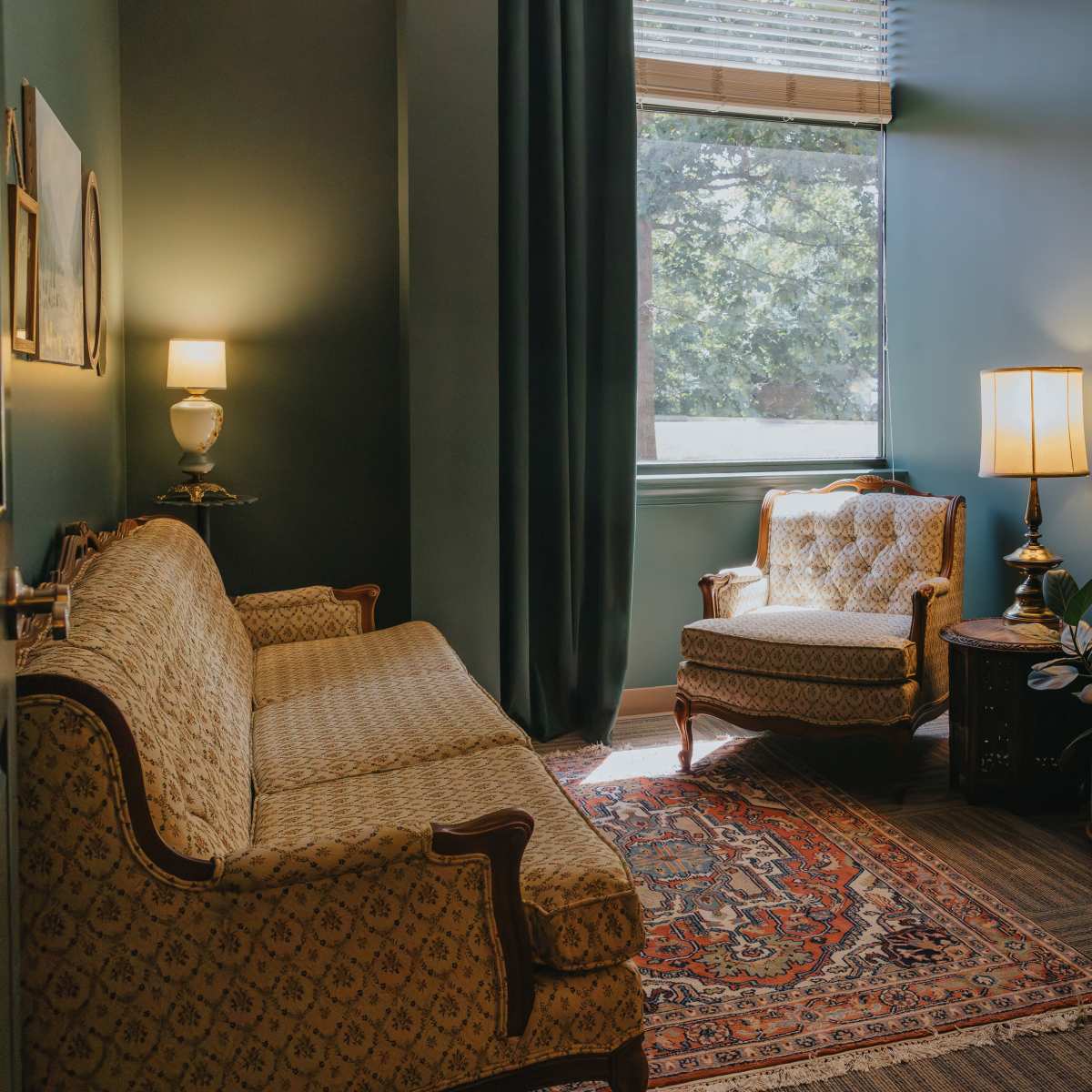 A cozy room featuring a patterned sofa, a vintage armchair, a wooden desk, and a large window allowing natural light to illuminate the space.