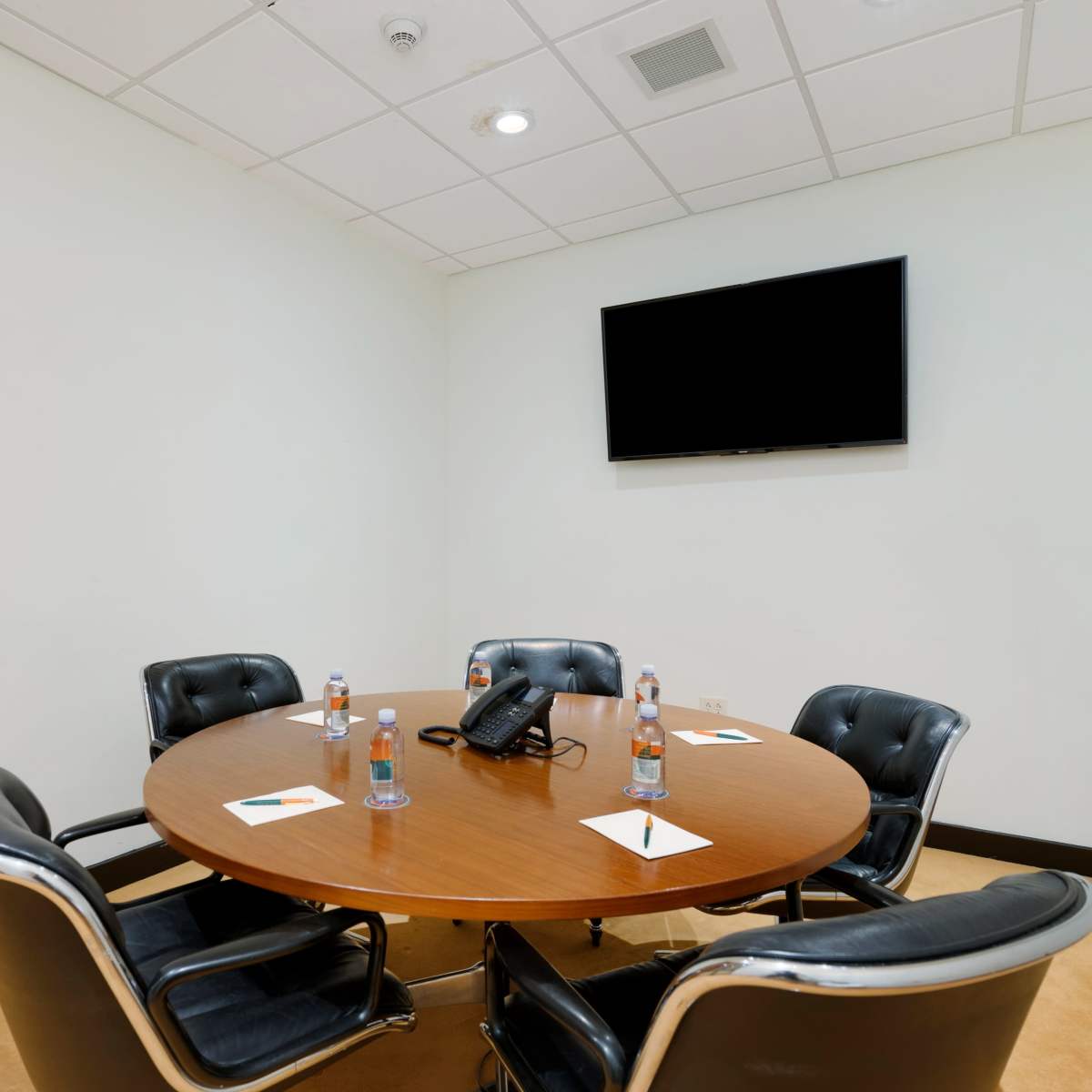 A round wooden conference table surrounded by five black leather chairs is set up in a meeting room, complete with a telephone and water bottles on the table, and a mounted television on the wall.