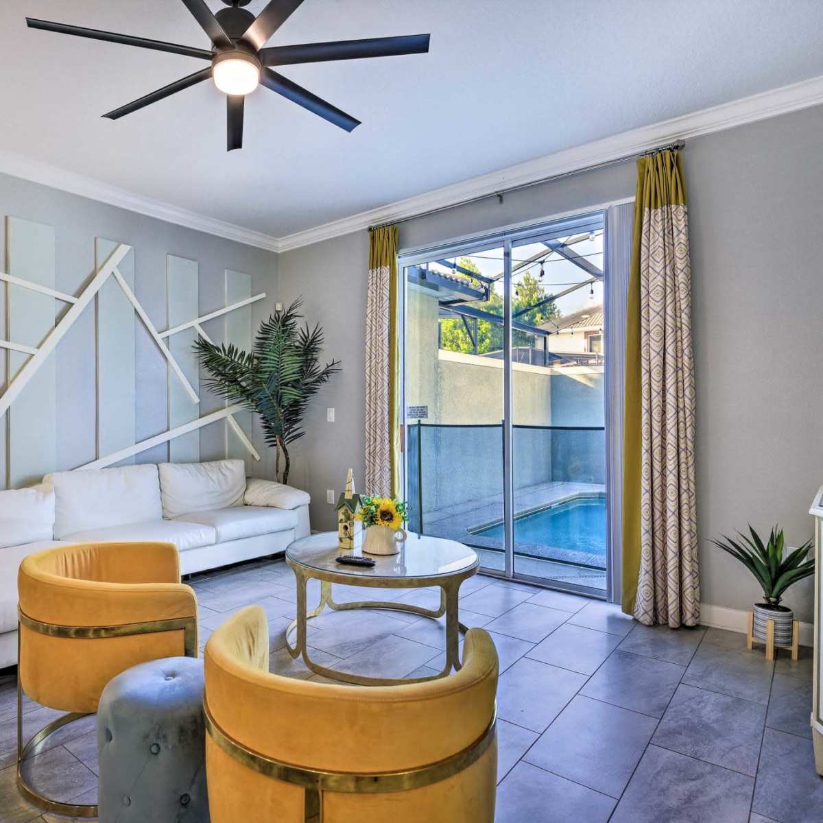 A modern living room features a white sectional sofa, yellow accent chairs, a round coffee table, and large windows overlooking a pool area.