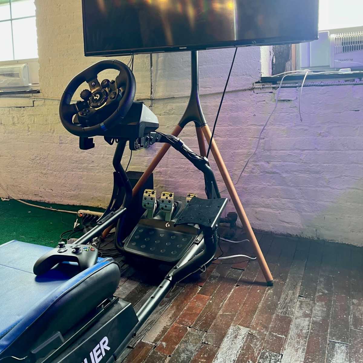 A racing simulation setup features a steering wheel and pedal system alongside an Xbox controller, positioned near a large flat-screen TV in a room with wooden flooring.