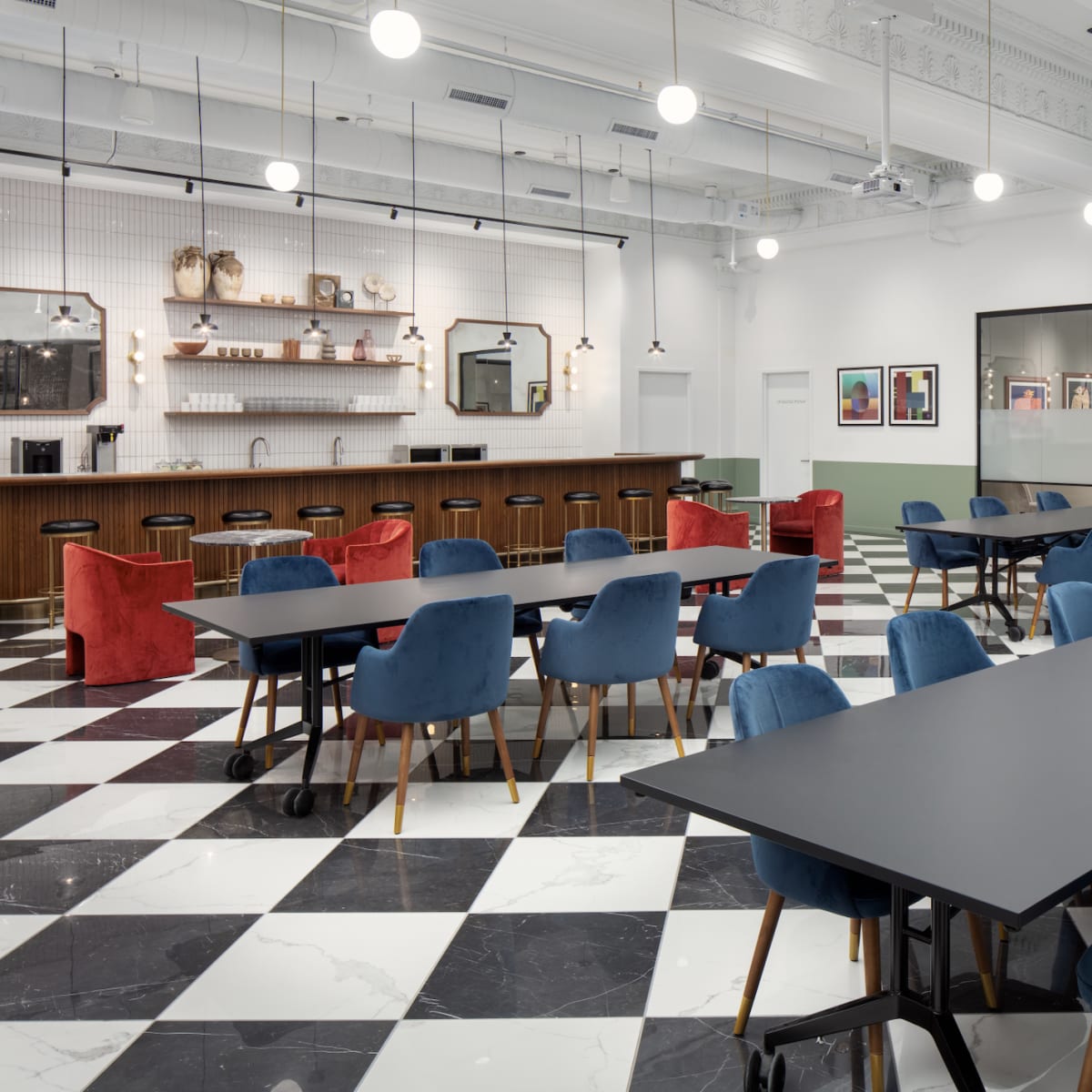 A modern, spacious dining area with checkered black and white flooring, featuring several tables surrounded by blue and red chairs, and a bar counter in the background.