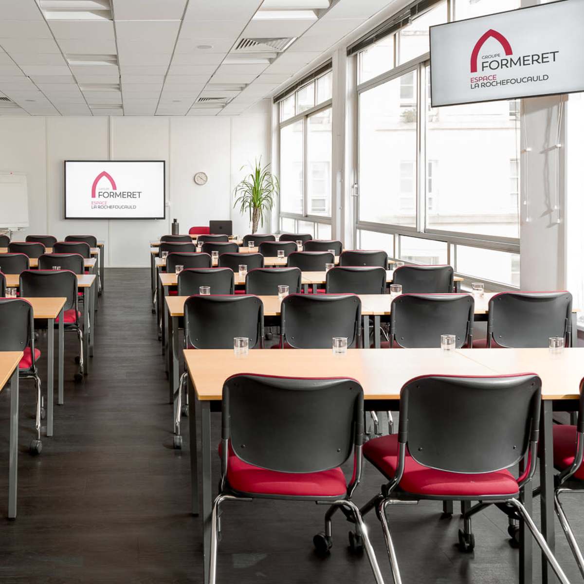 A spacious conference room with multiple rows of tables and chairs, equipped with two screens displaying a logo, and large windows allowing natural light.