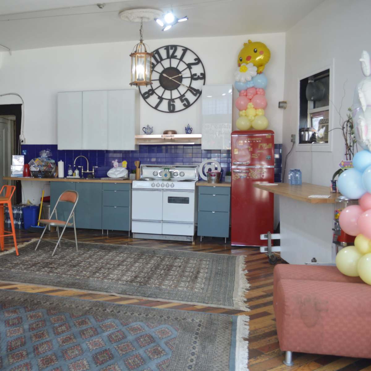 A café interior with a modern kitchen area, featuring a blue and gray color scheme, a vintage clock on the wall, and colorful decorations, including balloons.