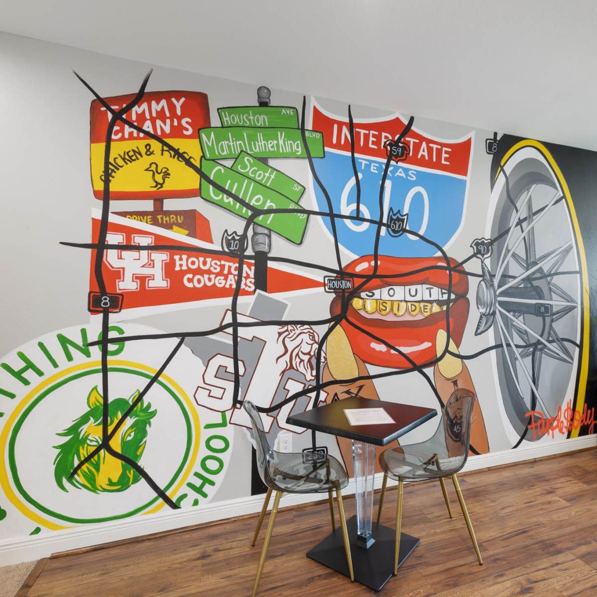 A modern living space featuring a colorful mural on the wall and a small black table with two clear chairs.