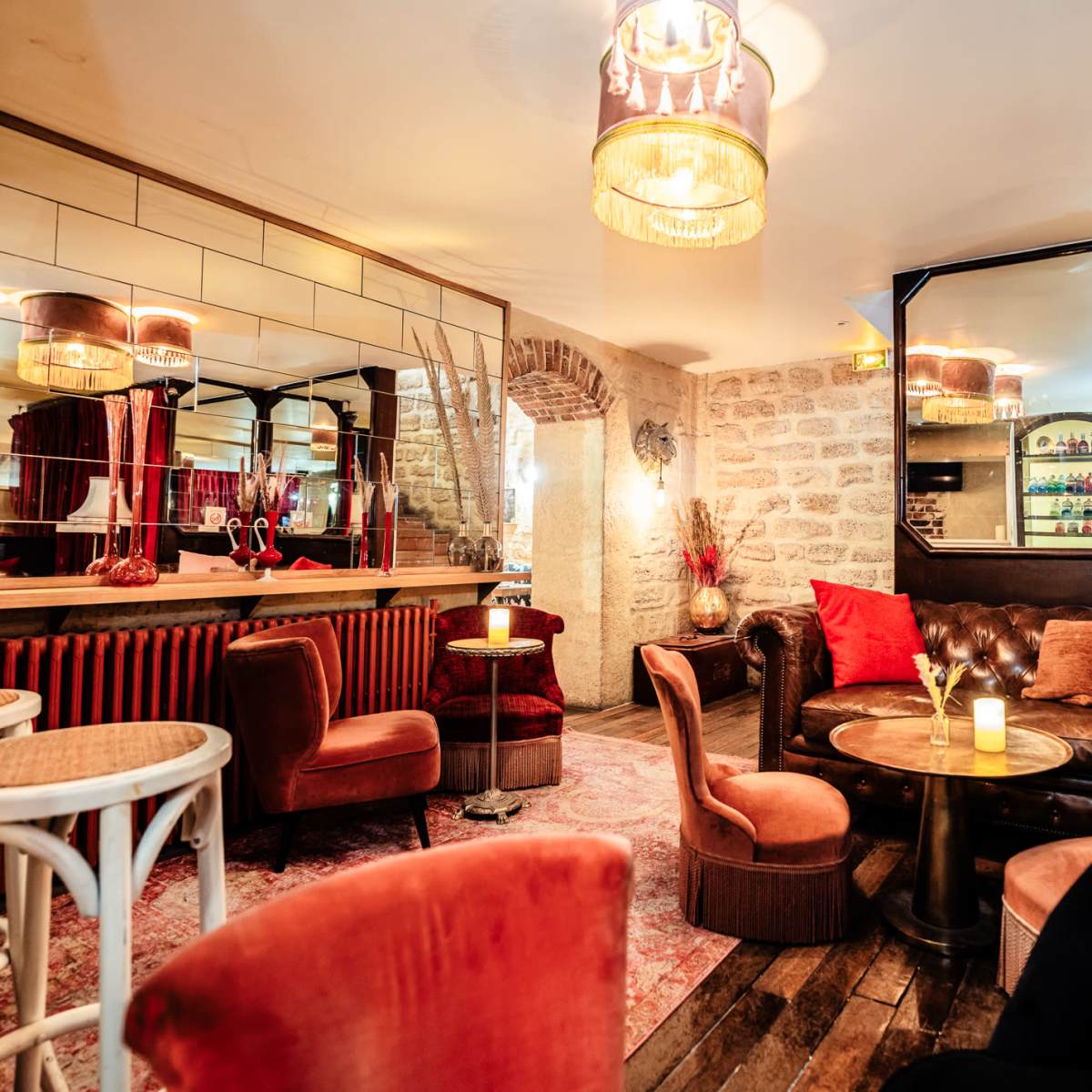 A stylish lounge area features a leather sofa, circular table, and illuminated chandeliers, with mirrors and red drapes adding to the ambiance.
