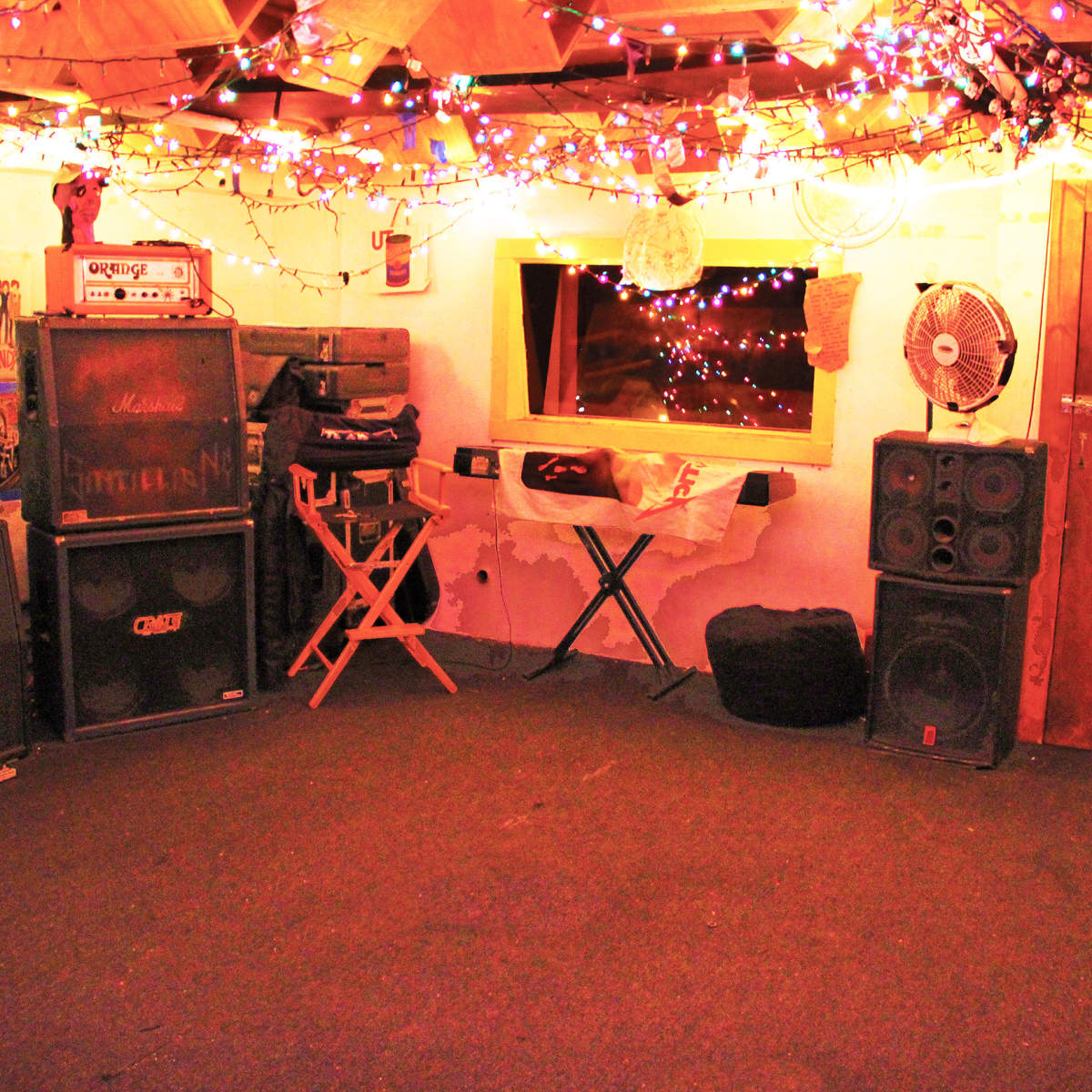 A dimly lit music practice room features amplifiers, speakers, and colorful string lights overhead.