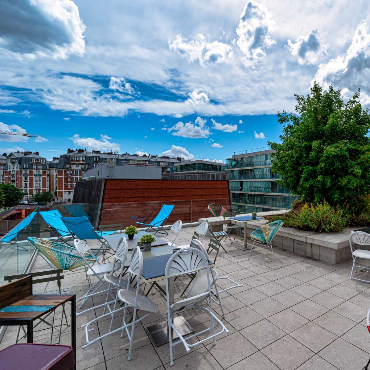 The rooftop terrace features a variety of tables and chairs surrounded by greenery under a cloudy sky.