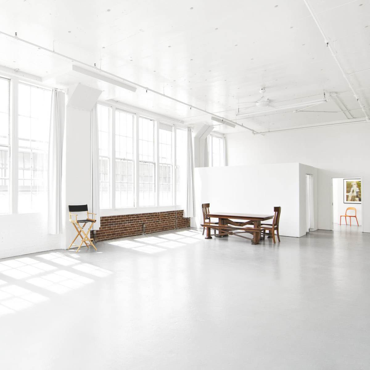 A bright, spacious room with large windows, a wooden table, a director's chair, and a white wall.