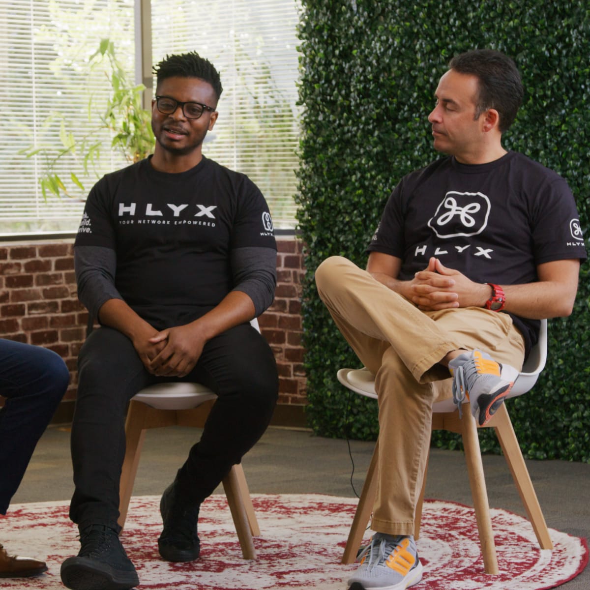 Three men sit on chairs in a well-lit room, wearing matching branded T-shirts, engaged in conversation.