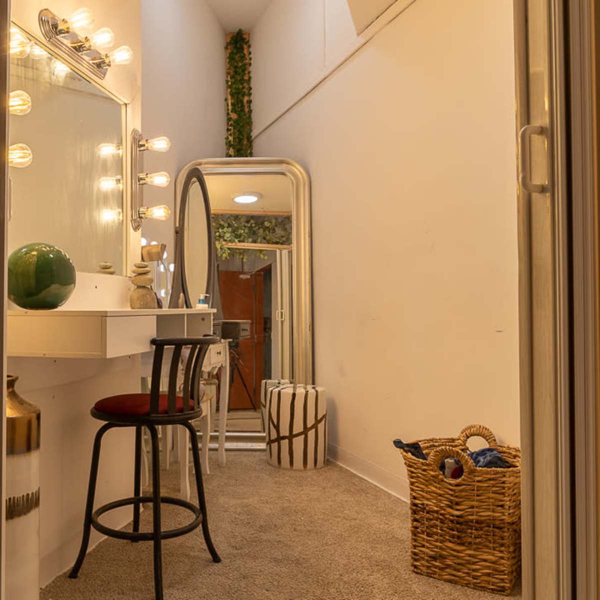 A well-lit dressing area featuring a vanity with lights, a mirror, a chair, and decorative items in a cozy hallway.