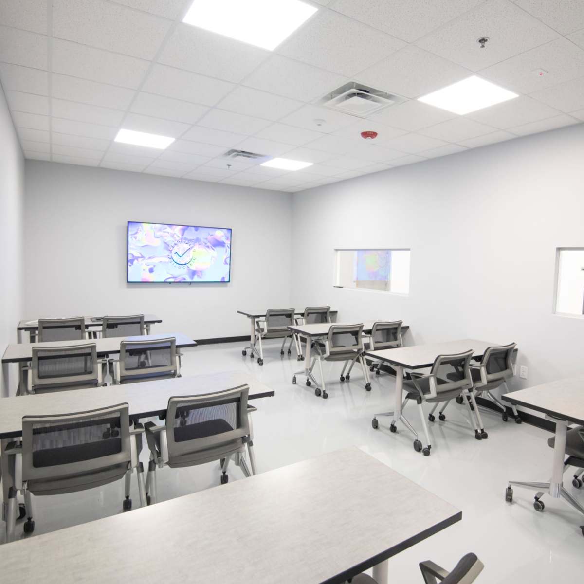 A modern classroom with multiple tables and chairs arranged in rows, featuring a large screen at the front and windows on one wall.
