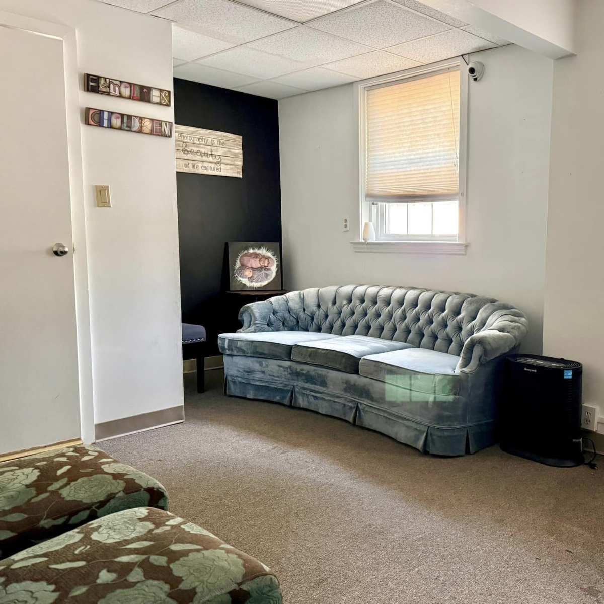 A room with a tufted blue couch, a small table on the left wall, and a door leading outside.