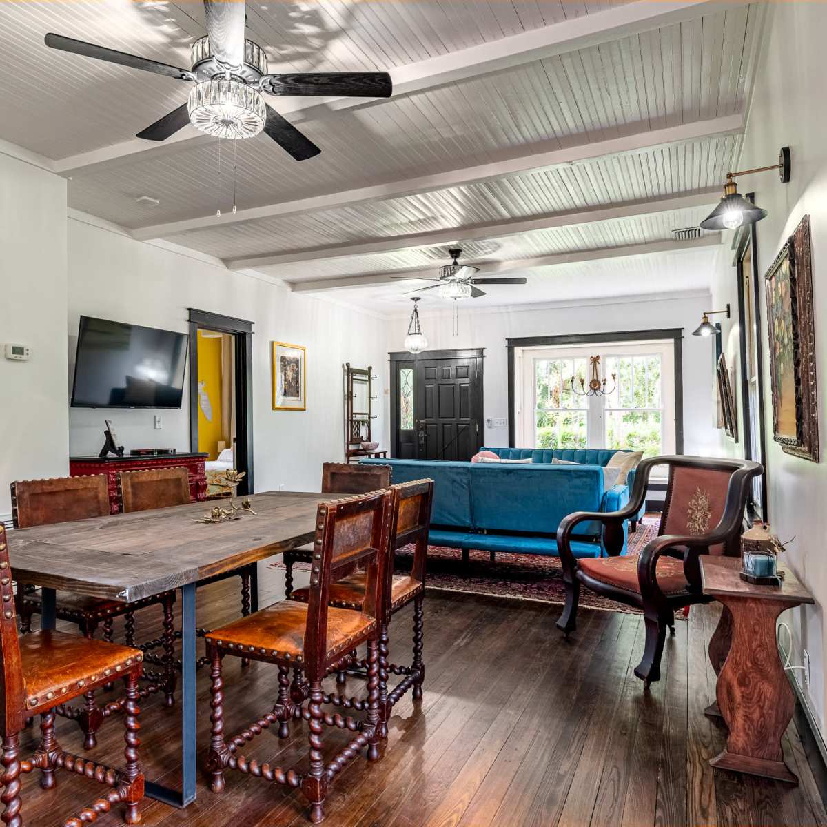 A living and dining area featuring a wooden dining table with chairs, a blue couch, and light fixtures, all under a ceiling with exposed beams.